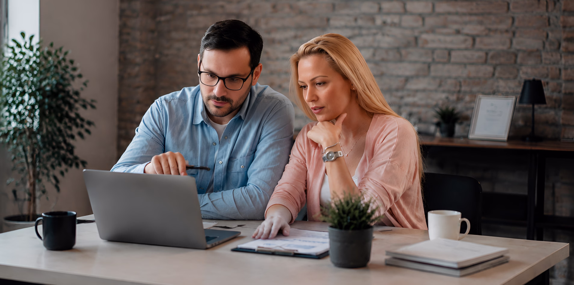 Two colleagues reviewing financial figures on a laptop in a brick-walled office