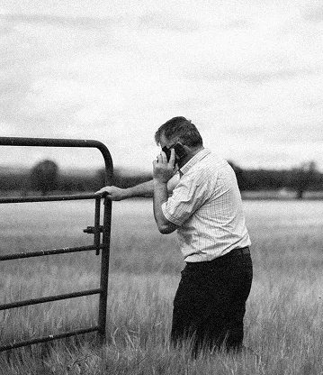 Man standing in a grassy field holding a metal gate with one hand and talking on a mobile phone with the other.