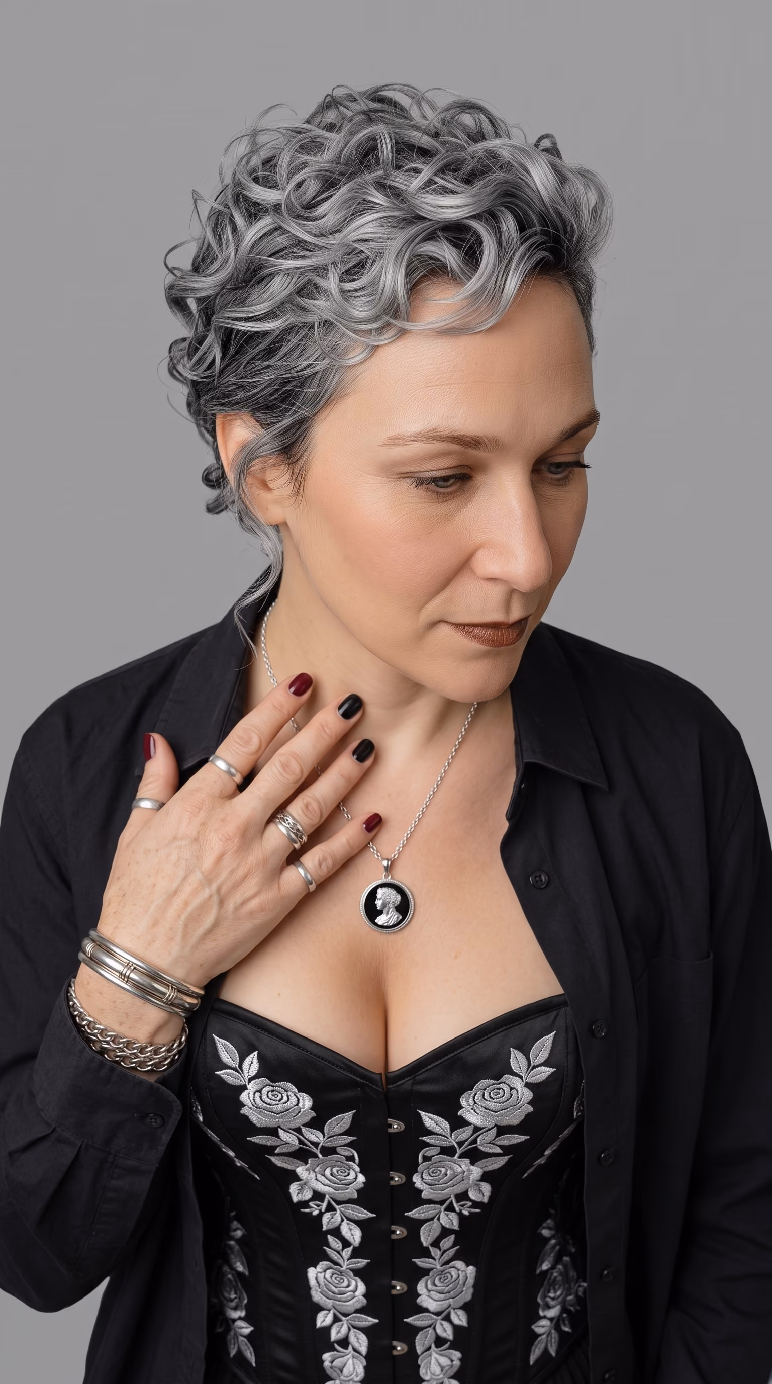 Woman with short curly gray hair wearing a black embroidered corset and black buttoned shirt, accessorized with multiple silver rings and bracelets, and a necklace with a black pendant depicting a classical profile, looking down with a serene expression.