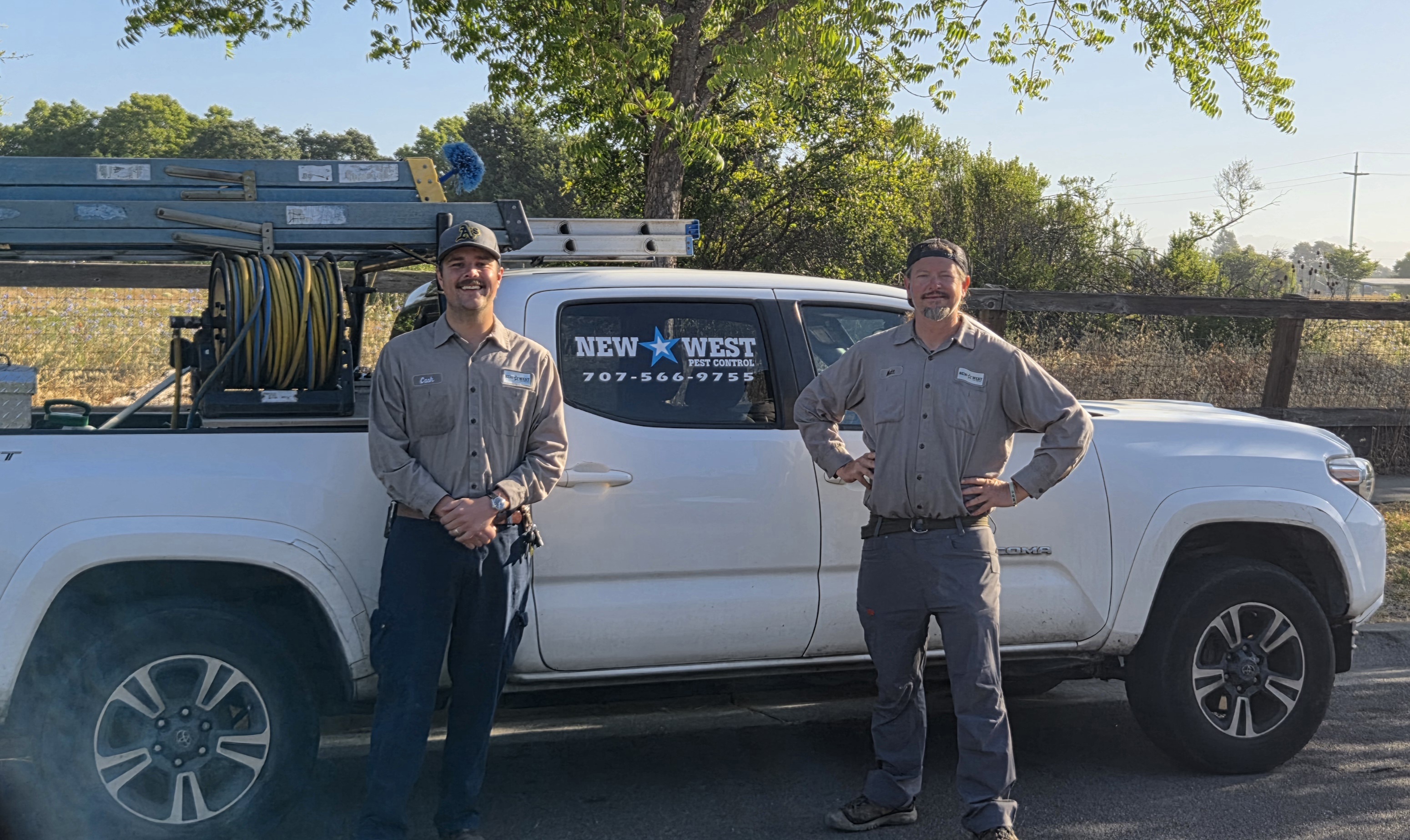Two pest control workers stand in front of a white truck with ladders and hoses in the bed.