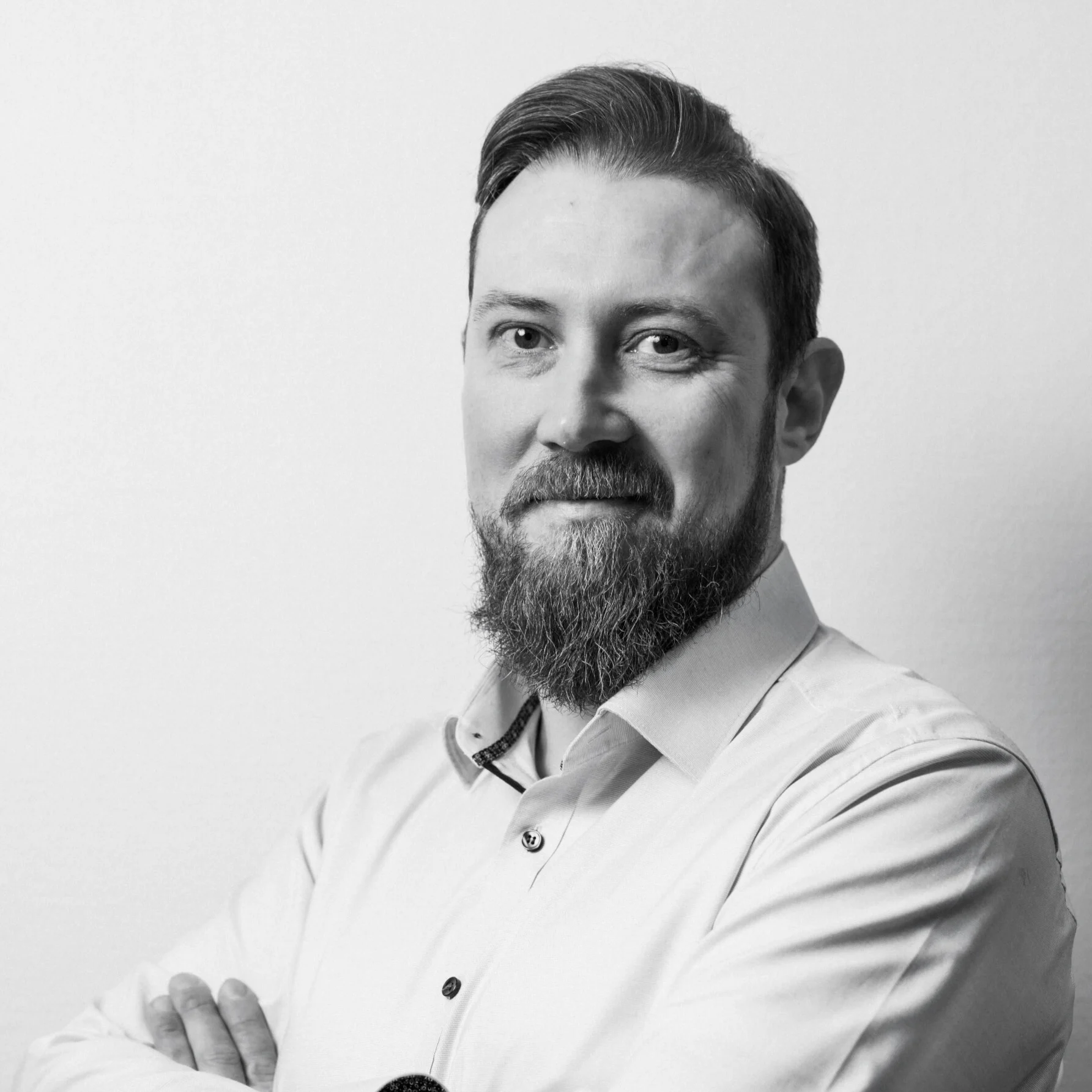 Black and white portrait of a smiling man with a full beard and neatly combed hair wearing a buttoned light shirt with arms crossed.