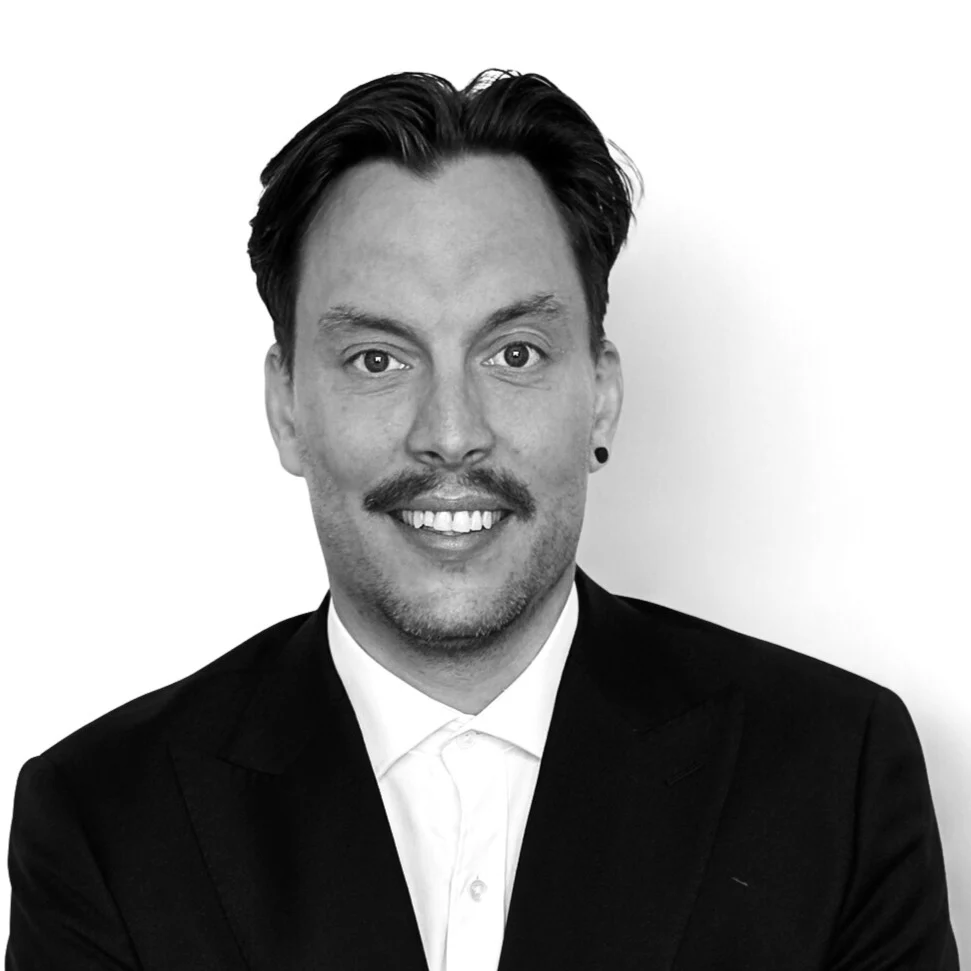 Black and white portrait of a smiling man with dark hair and a mustache, wearing a dark suit jacket over a white collared shirt, with a small earring in his left ear, against a plain light background.