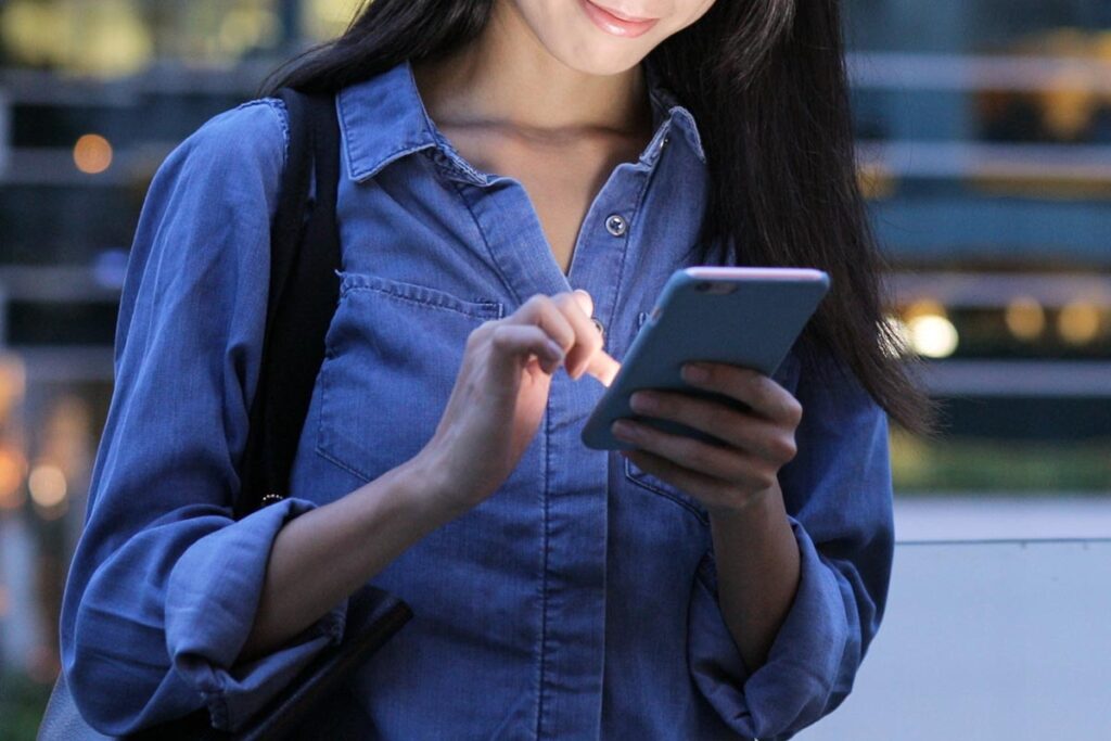 A photo of a woman with a backpack in a denim shirt typing on a smartphone.