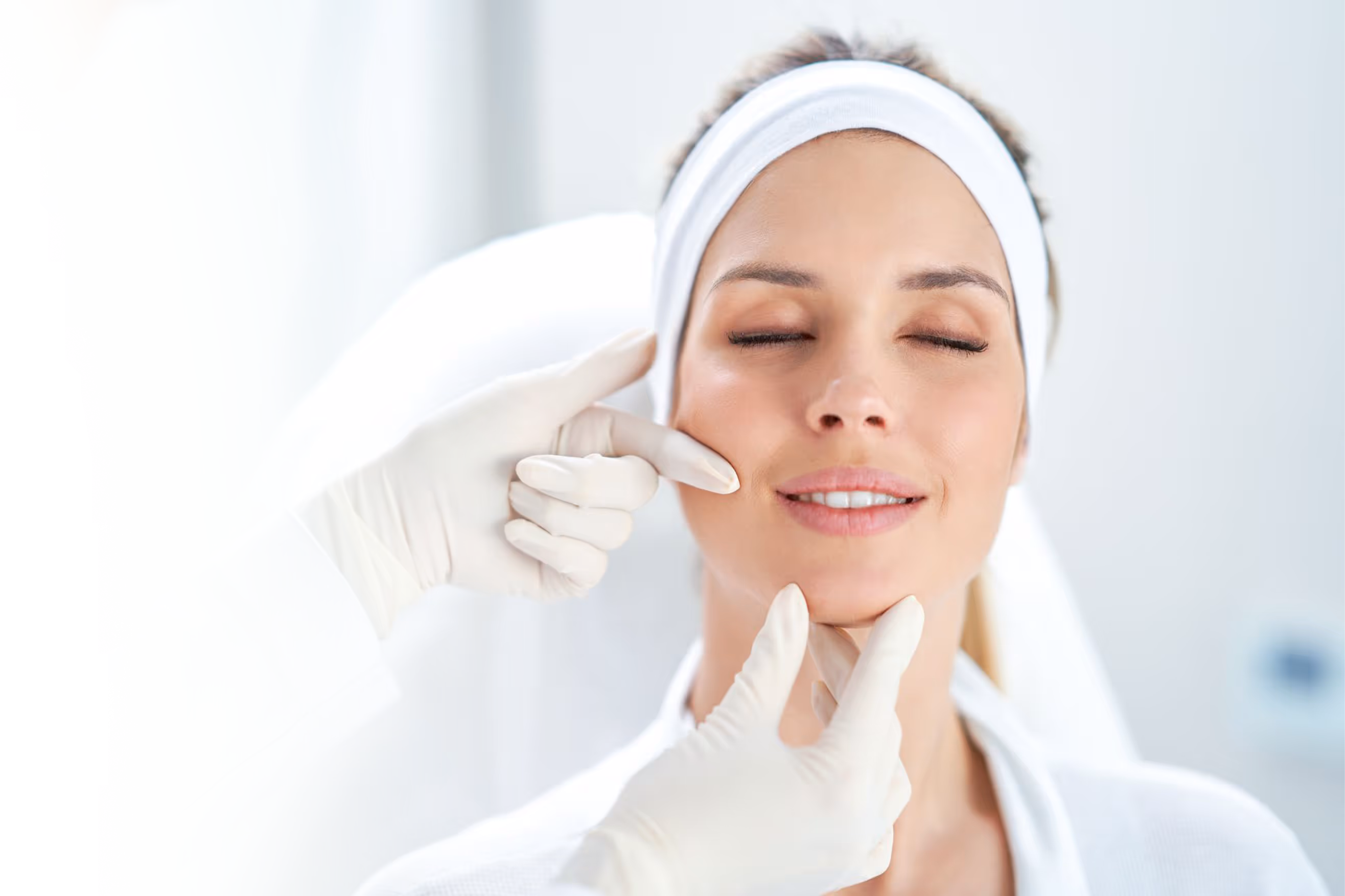 Woman with a headband receiving a facial skin check from a person in gloves in a clinical setting.