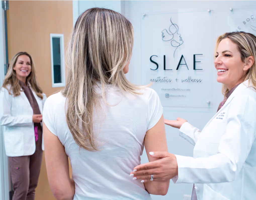 Two women in white coats greeting and talking with a patient in a wellness clinic lobby.