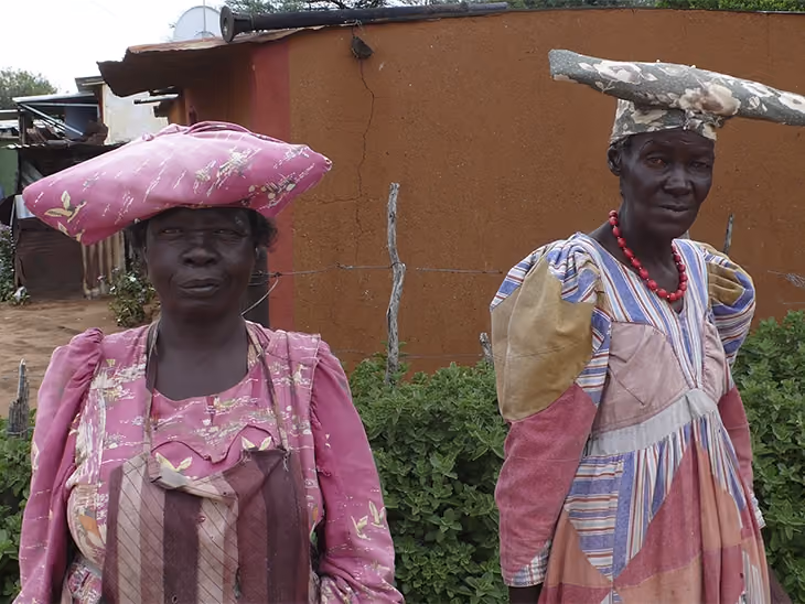 Zwei afrikanische Frauen in bunter traditioneller Kleidung und auffälligen Hüten vor einem Gebäude.