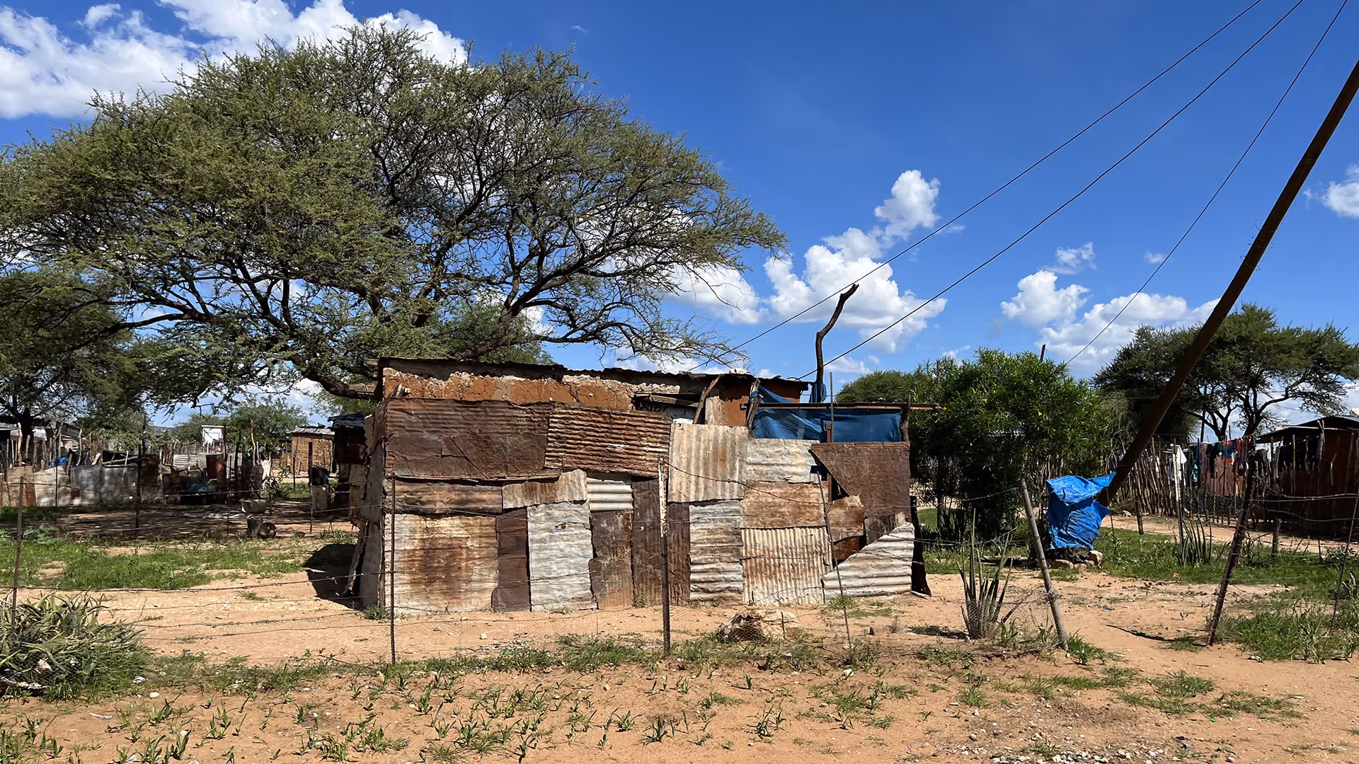Well-weathered shack made of corrugated metal sheets under trees with blue sky and scattered clouds.