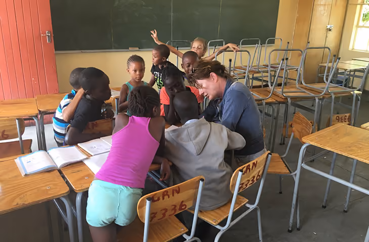 Lehrerin umgeben von acht Kindern an Tischen in einem Klassenzimmer mit Tafel im Hintergrund.