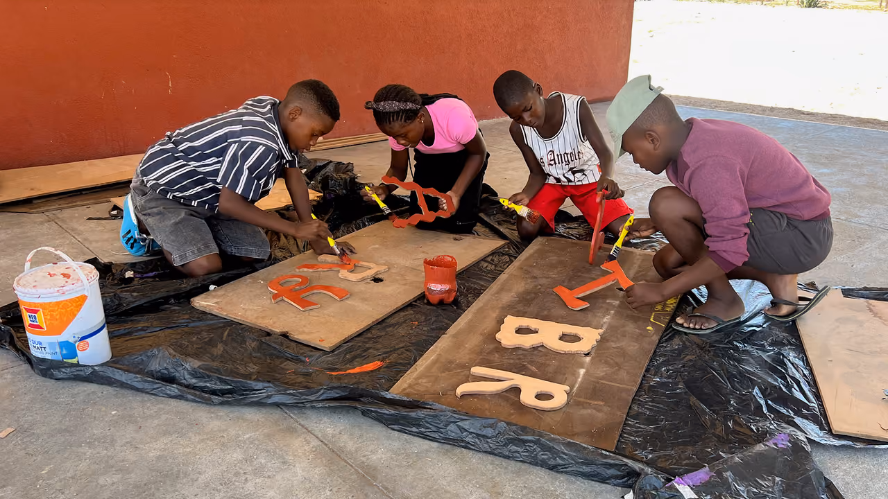 Vier Kinder malen Buchstaben mit orangener Farbe auf Holzplatten, sitzend auf dem Boden.