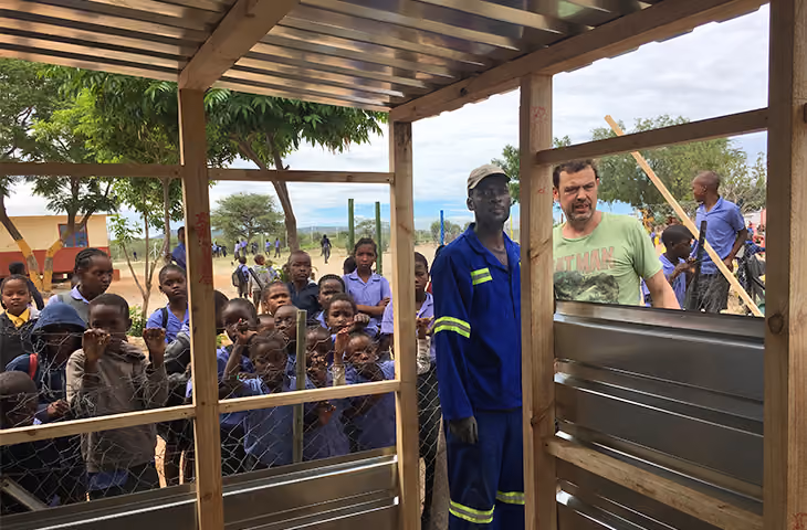 Eine Gruppe Schulkinder steht hinter Zaun, zwei Männer in eines überdachten Holzrahmens im Freien.