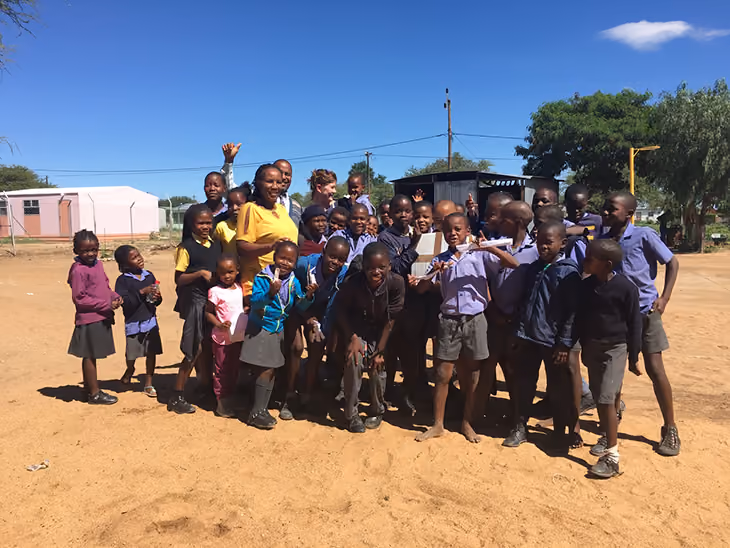 Gruppe von Schulkindern und Lehrerin lachen zusammen auf einem sonnigen Schulhof mit blauem Himmel.