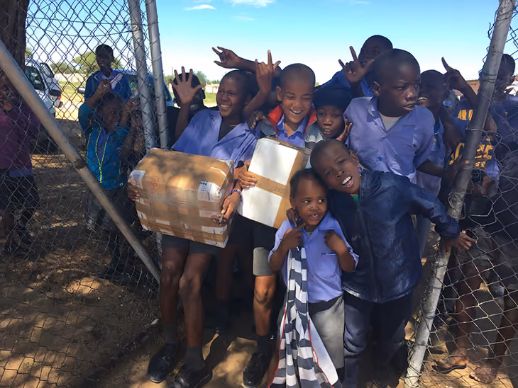 Group of smiling children in school uniforms holding packages and standing by a chain-link fence.