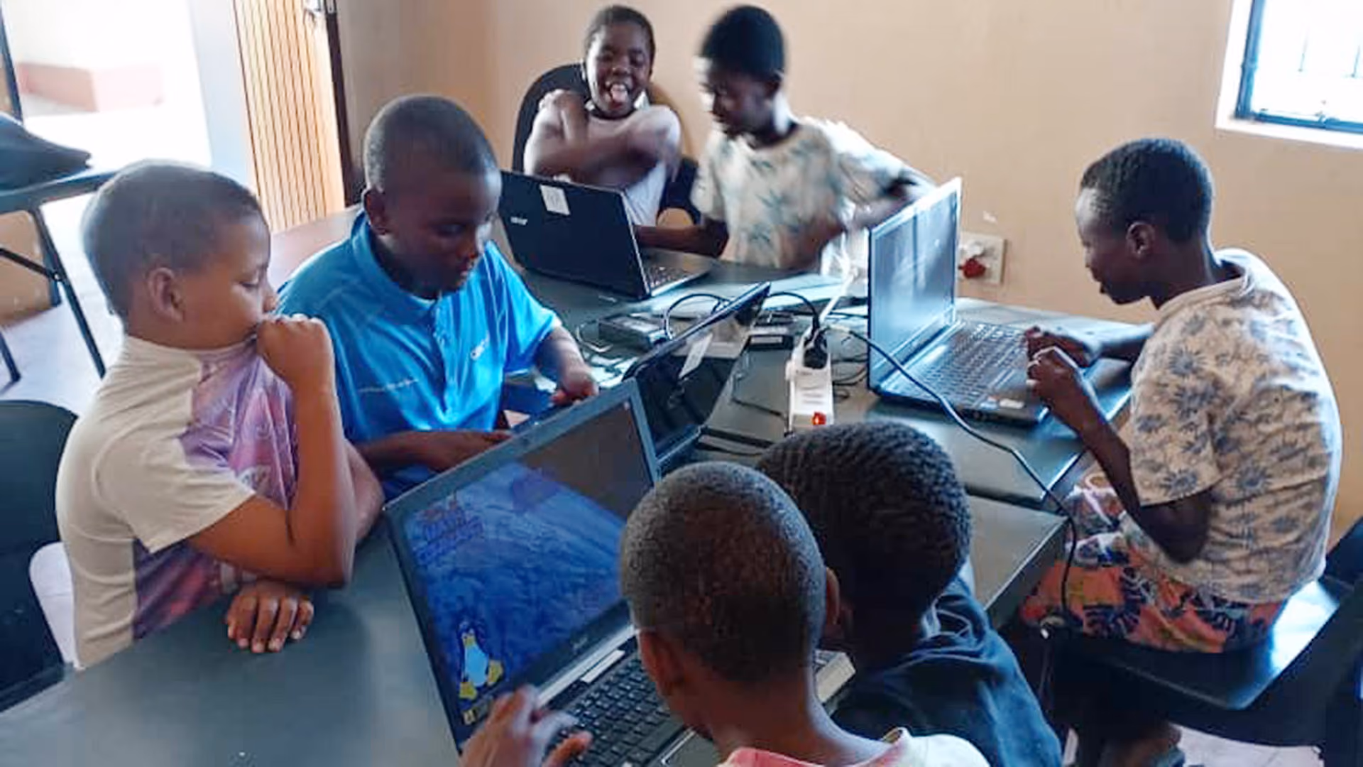 Sechs Kinder sitzen um Tische und arbeiten gemeinsam an Laptops in einem Klassenzimmer.