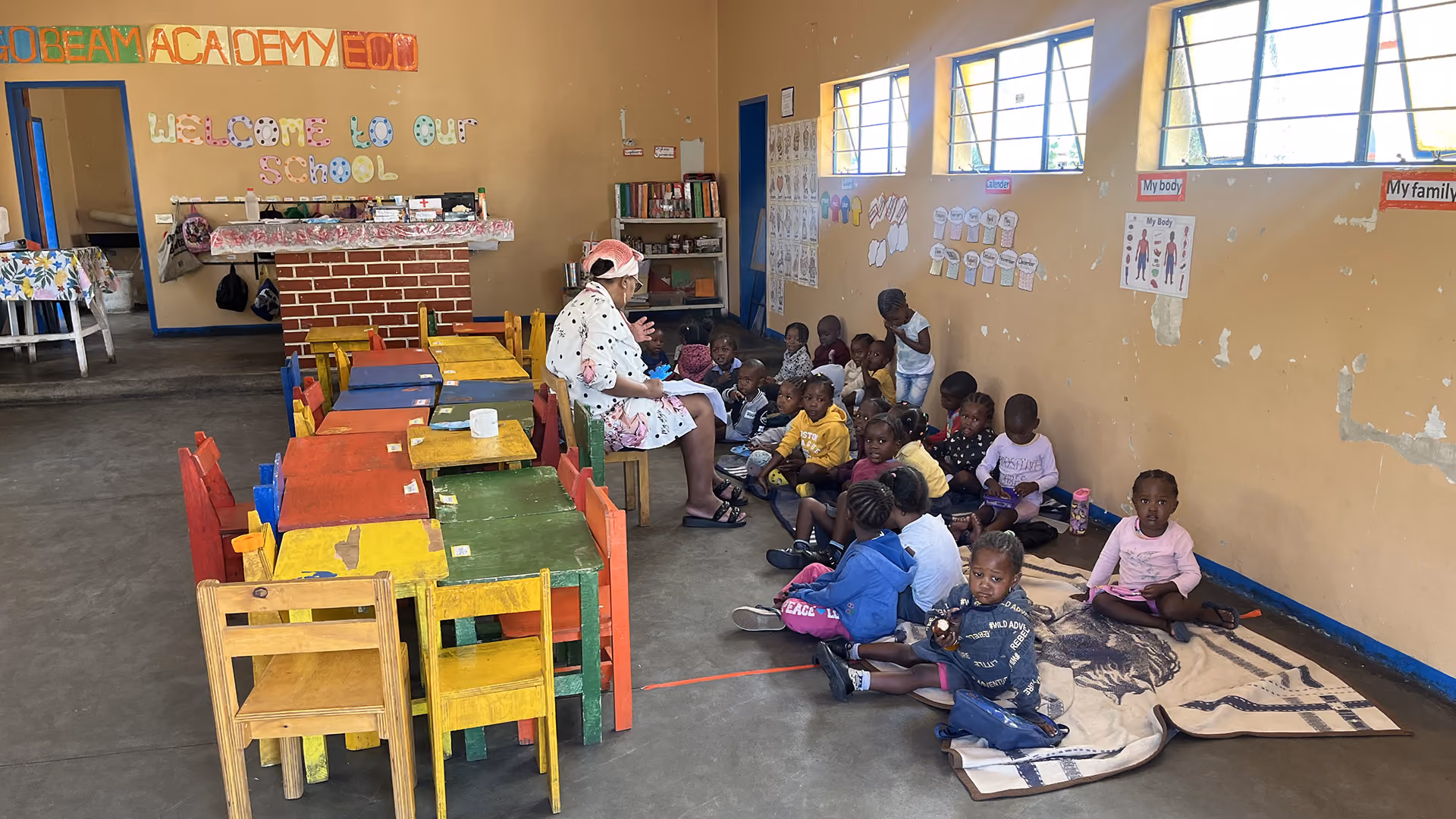 Lehrerin liest Kindern in einem Klassenraum mit bunten Tischen und Stühlen vor.