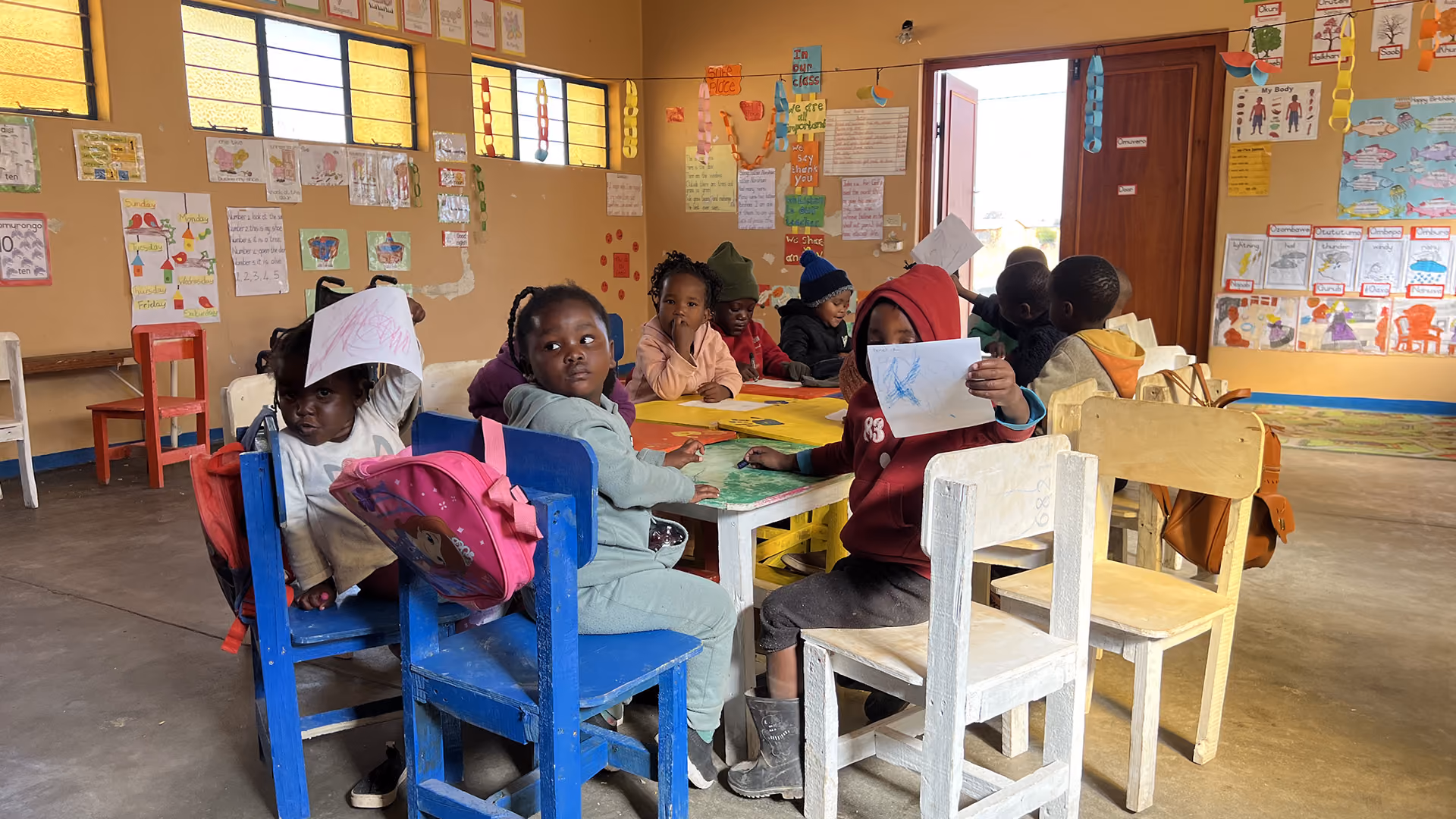 Kinder sitzen an Tischen in einem Klassenzimmer mit bunten Zeichnungen und Postern an den Wänden.