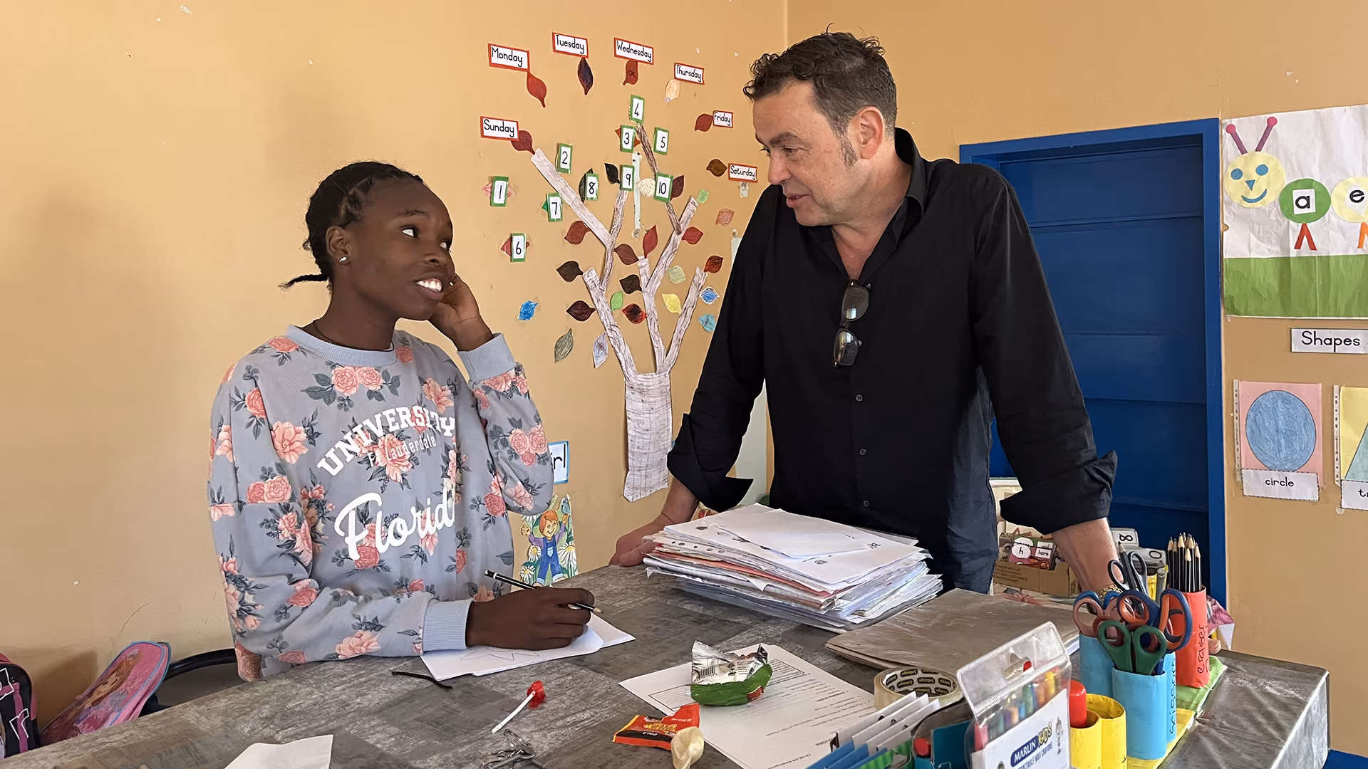 Lehrerin spricht mit Schülerin in Klassenzimmer mit Lernposter und Bastelmaterialien.