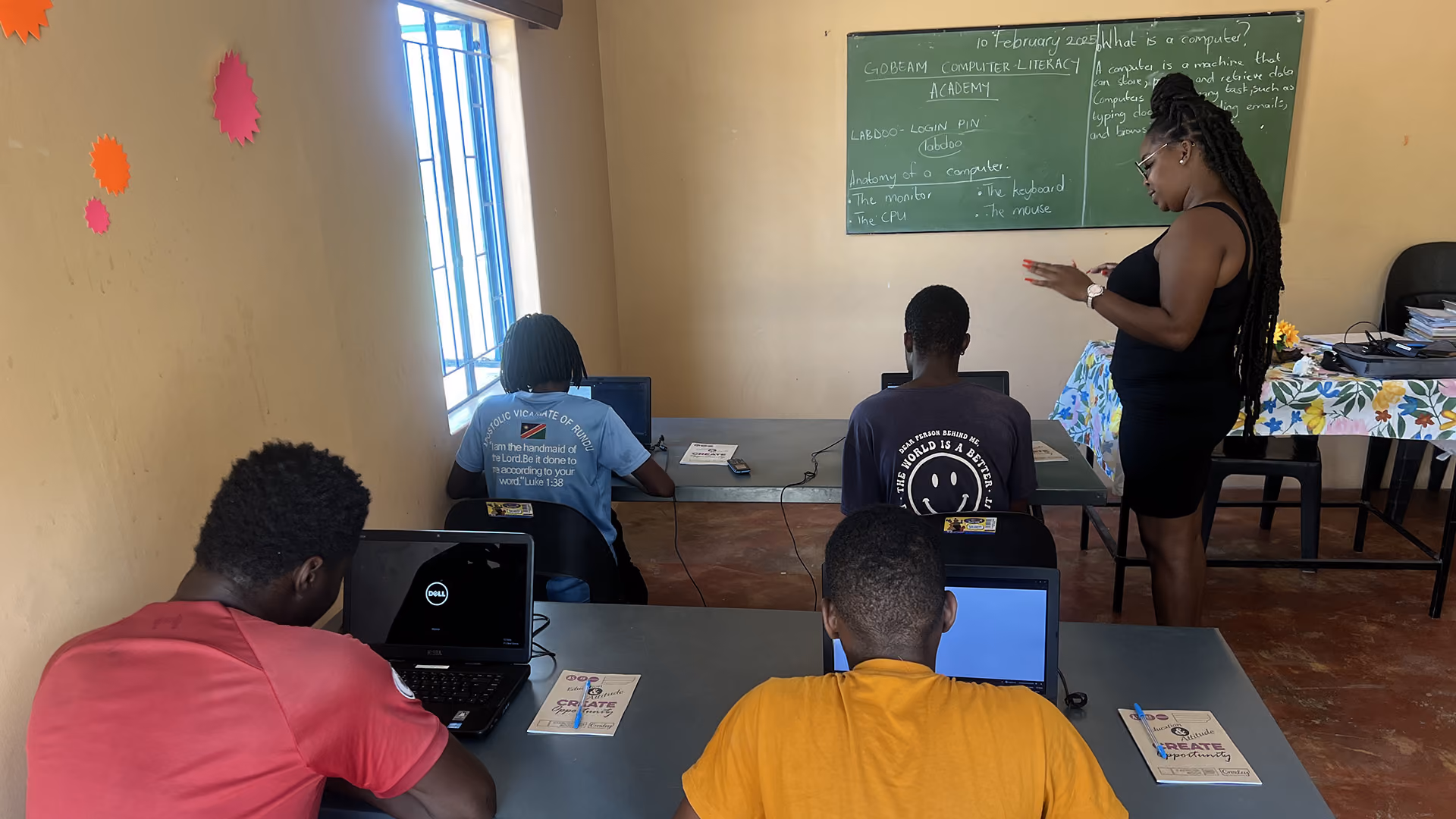 Vier Schüler an Laptops in einem Klassenzimmer, während eine Lehrerin vorne vor einer Tafel steht.