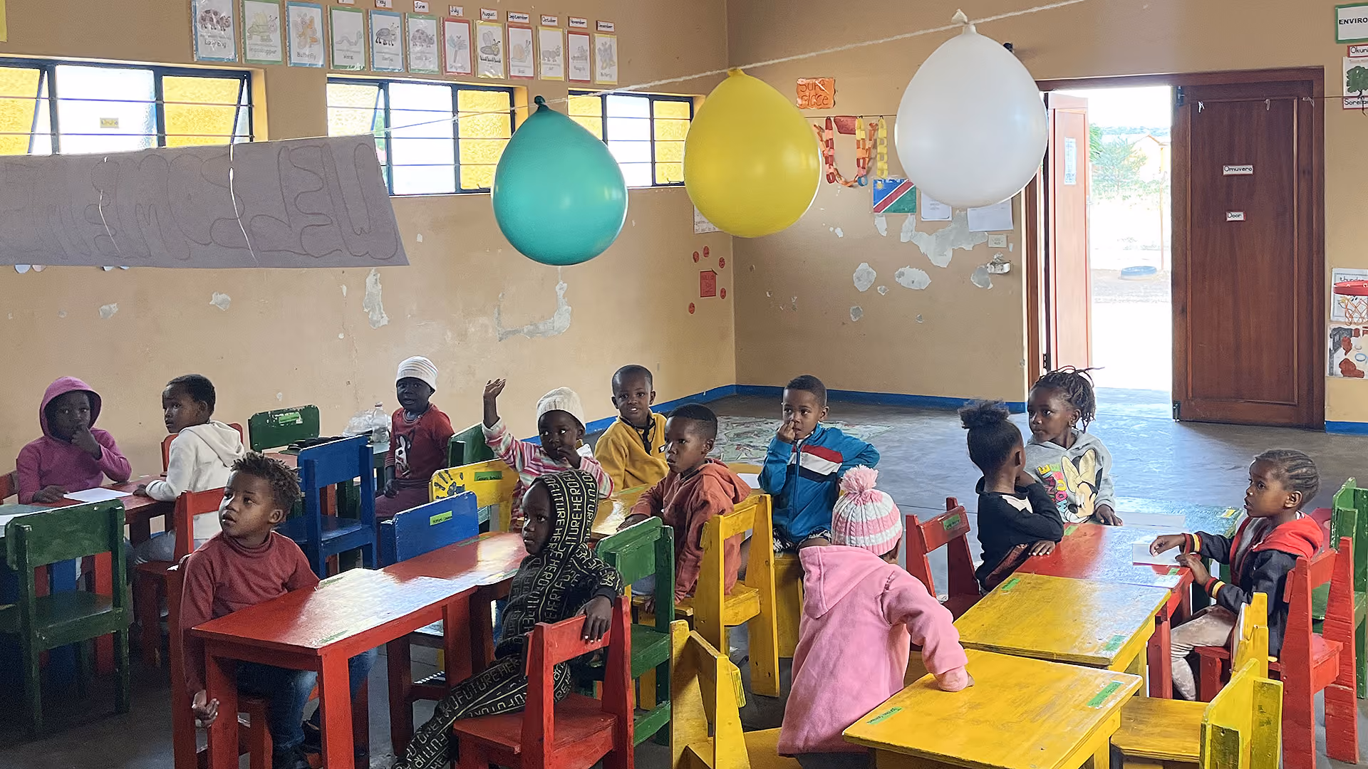 Vorschulkinder sitzen in einem Klassenzimmer an bunten Tischen unter Luftballons.