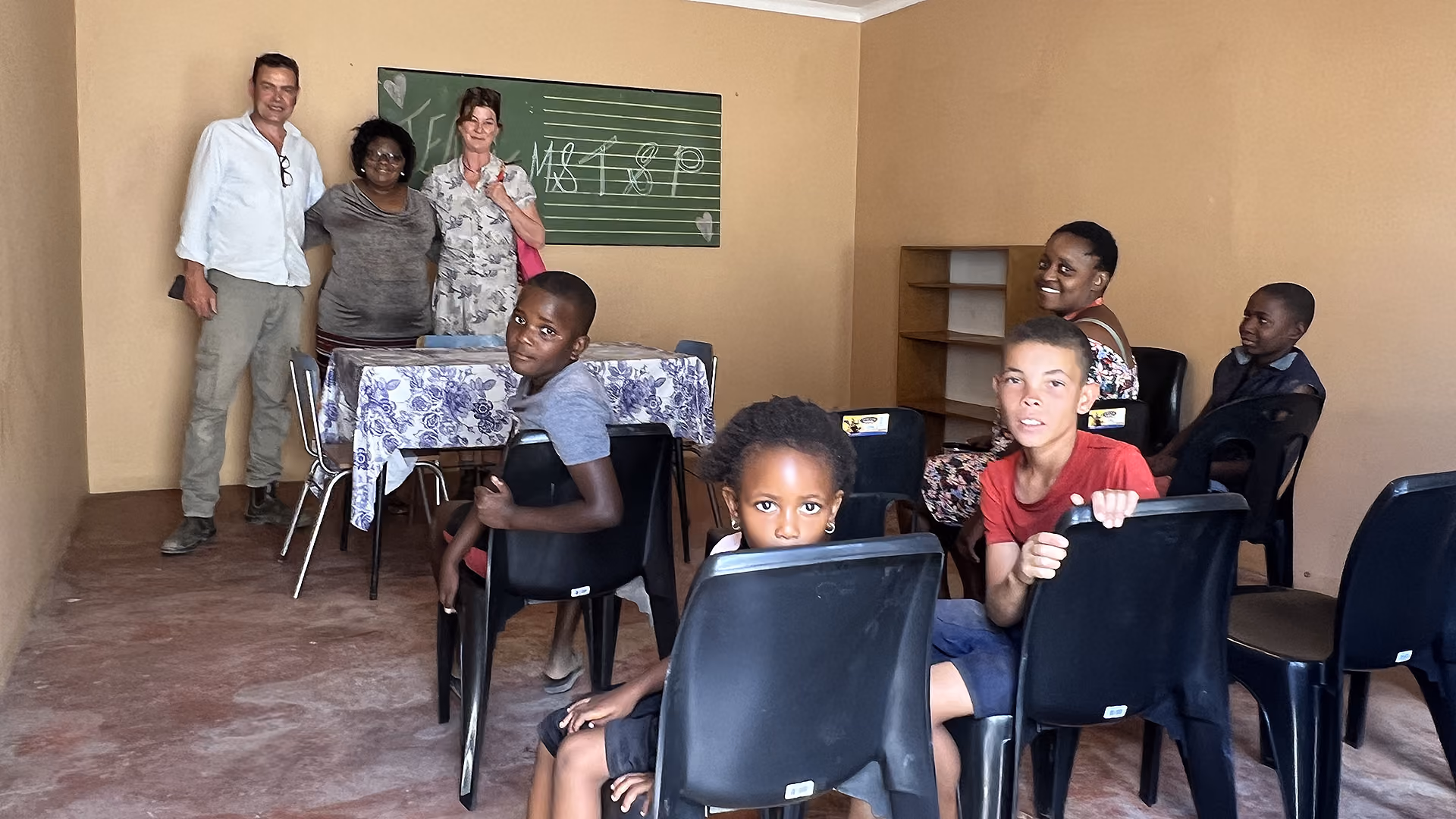 Drei Erwachsene und mehrere Kinder sitzen und stehen in einem einfachen Klassenzimmer mit leeren Stühlen.