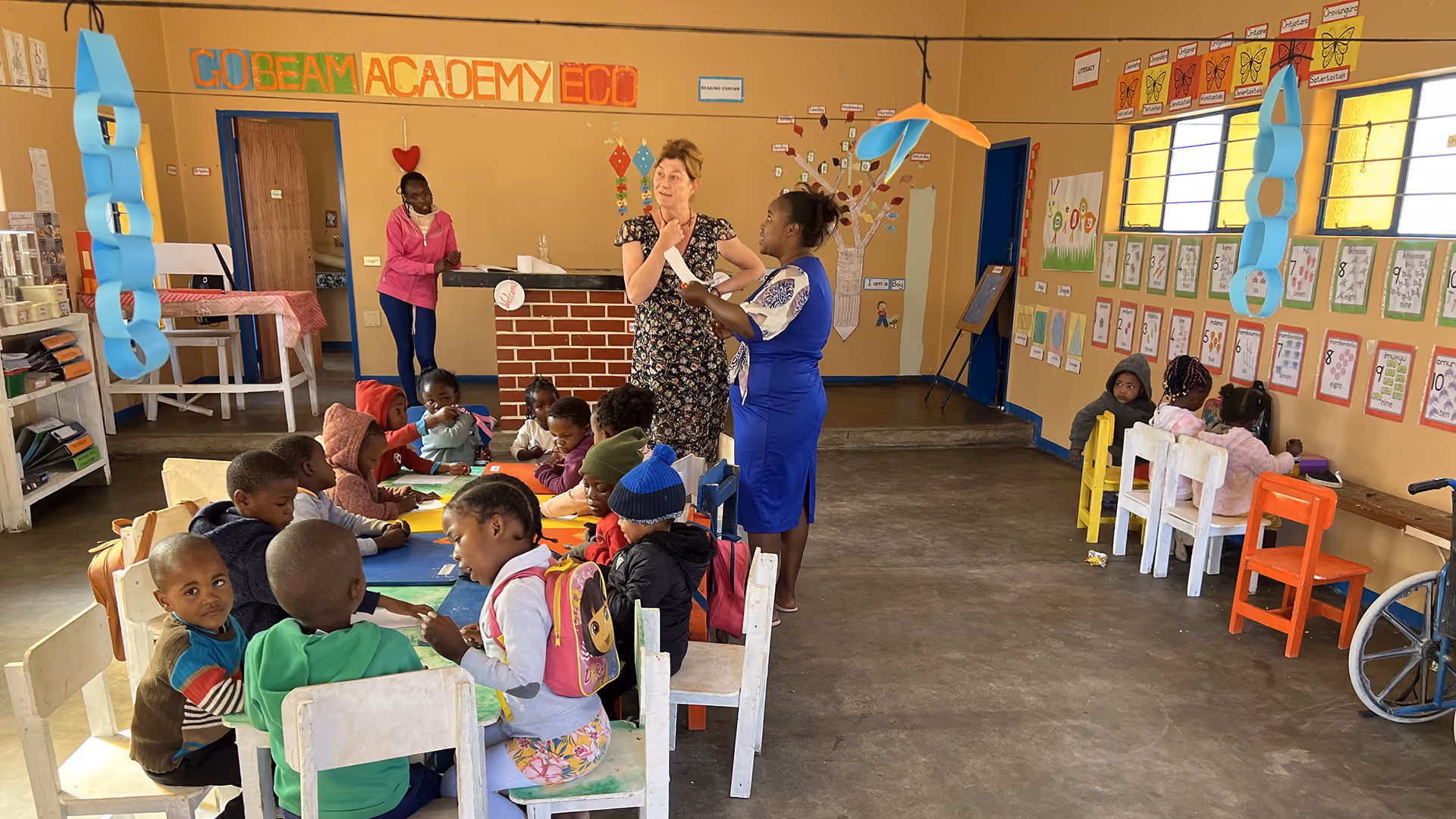 Kinder sitzen an Tischen in einem Klassenzimmer, während zwei Frauen vorne sprechen.
