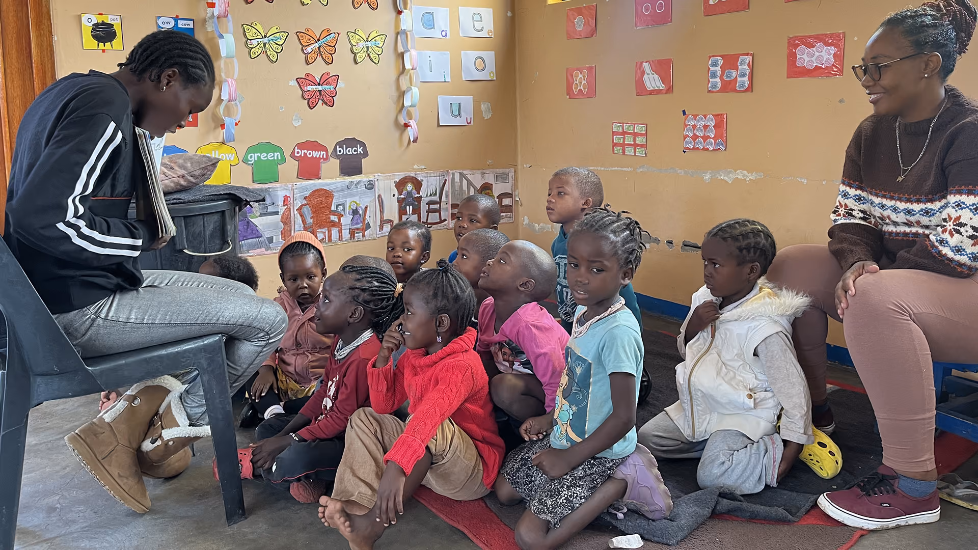 Kinder sitzen auf dem Boden im Klassenraum und hören einer vorlesenden Person aufmerksam zu.
