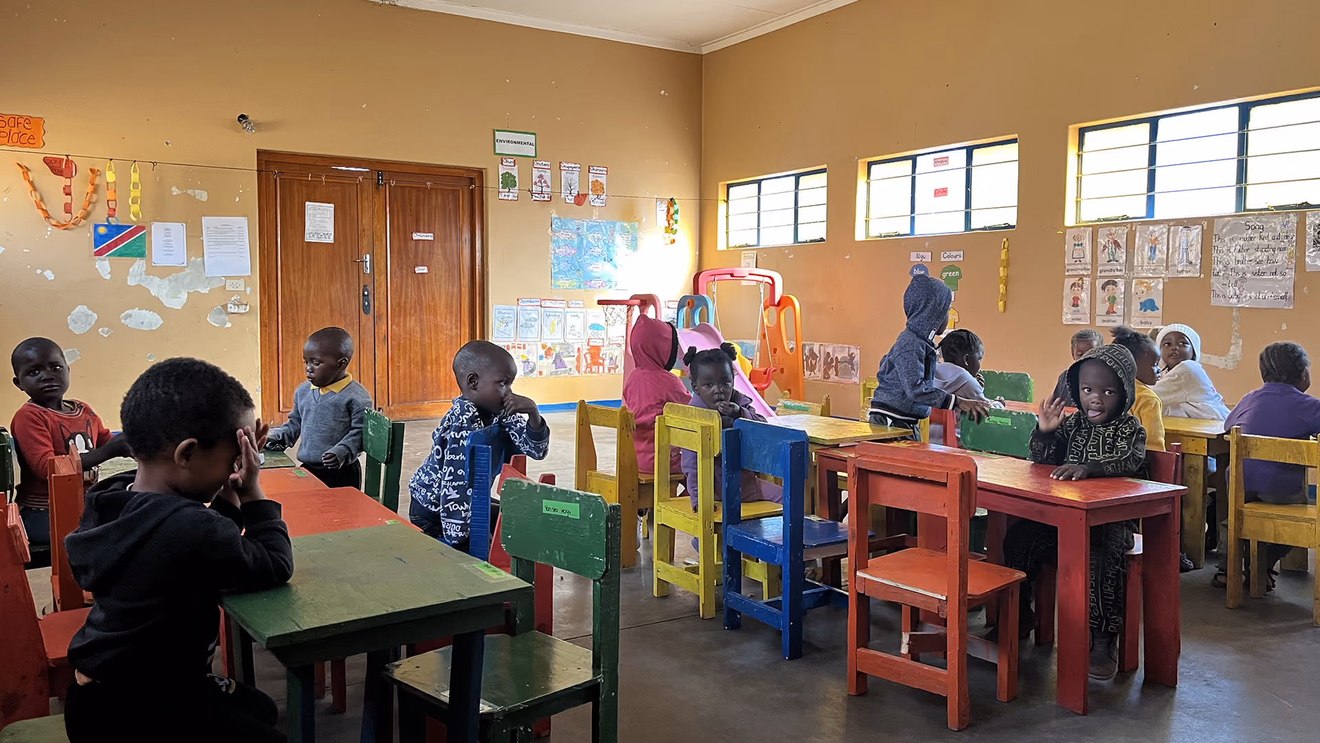 Kinder sitzen an bunten Tischen in einem Klassenzimmer mit Lernplakaten an den Wänden.