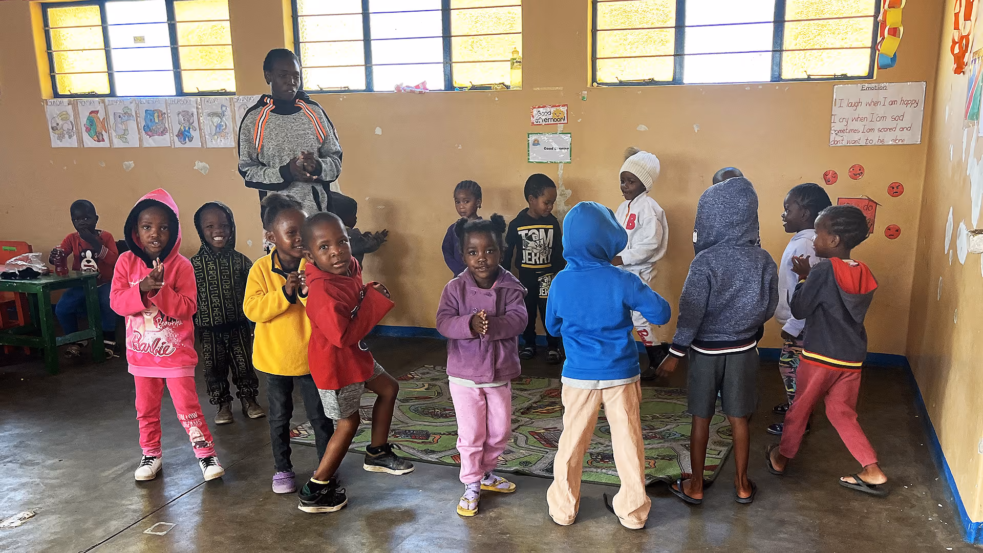 Kinder in warmen Kleidern stehen und klatschen in einem Klassenraum mit bunten Zeichnungen an der Wand.
