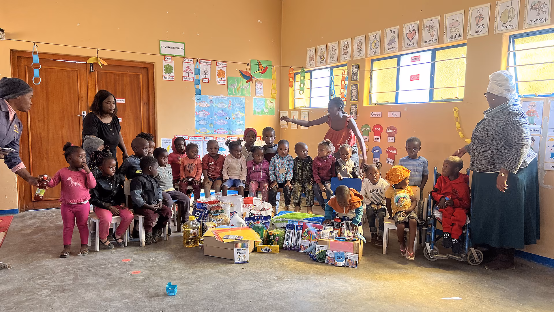 Kinder sitzen auf Stühlen in einem Klassenraum mit Lehrern und Schulsachen auf dem Boden.