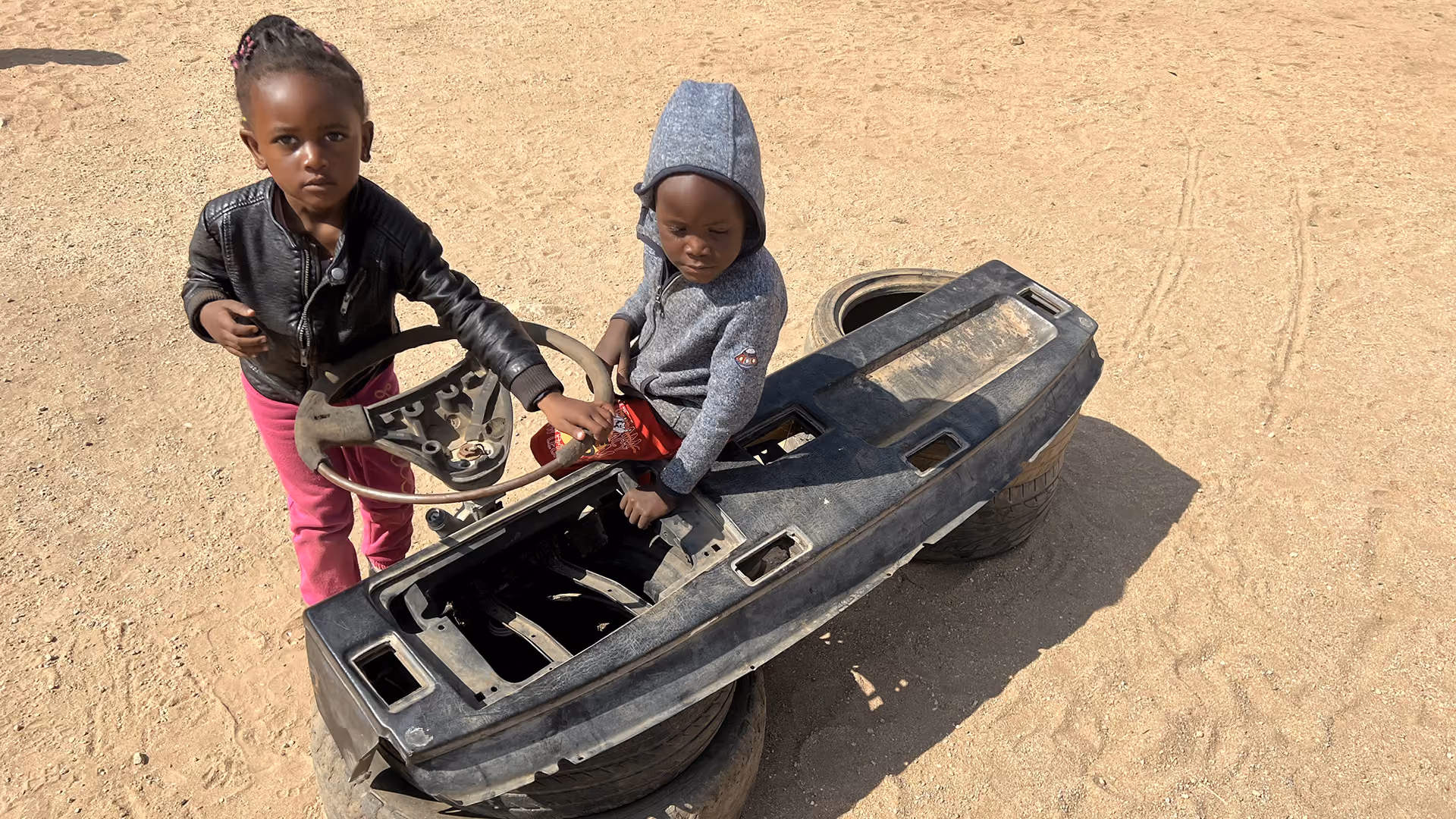 Zwei Kinder spielen draußen mit einem alten Autoteil und einem Lenkrad auf Sandboden.