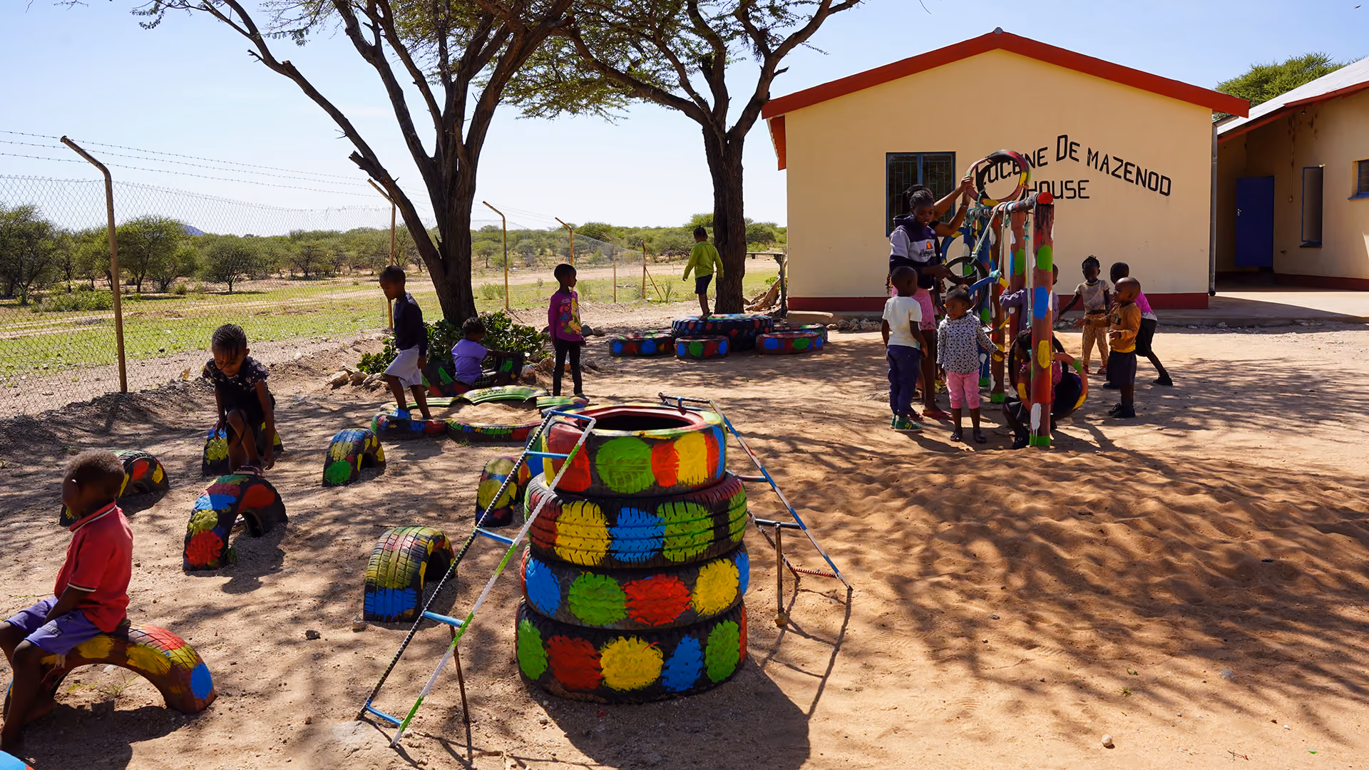 Kinder spielen draußen auf einem Spielplatz mit bunten Reifen und Spielgeräten vor einem Schulgebäude.