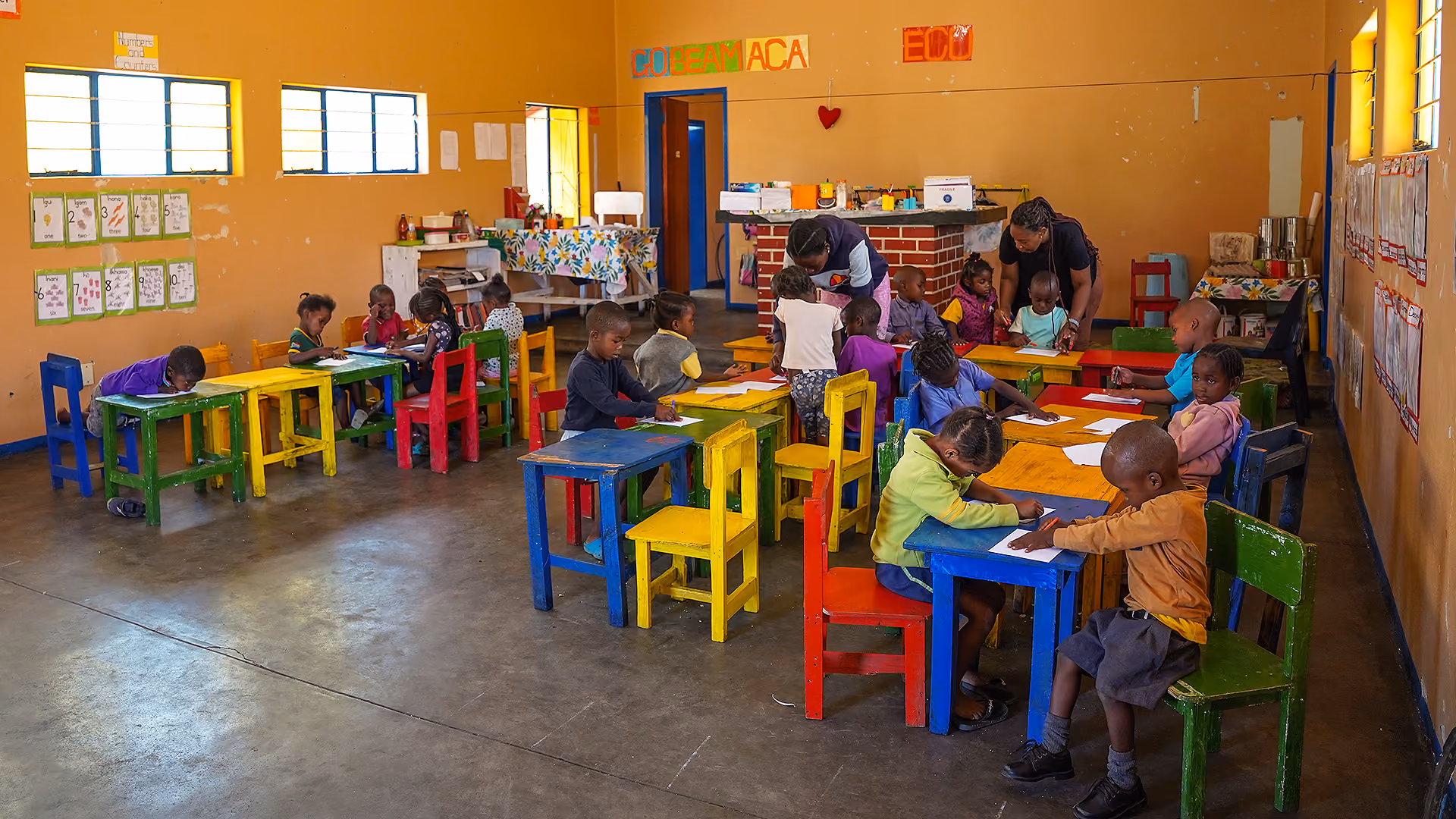 Kinder sitzen an bunten Tischen und Stühlen in einem Klassenraum mit zwei Lehrerinnen.