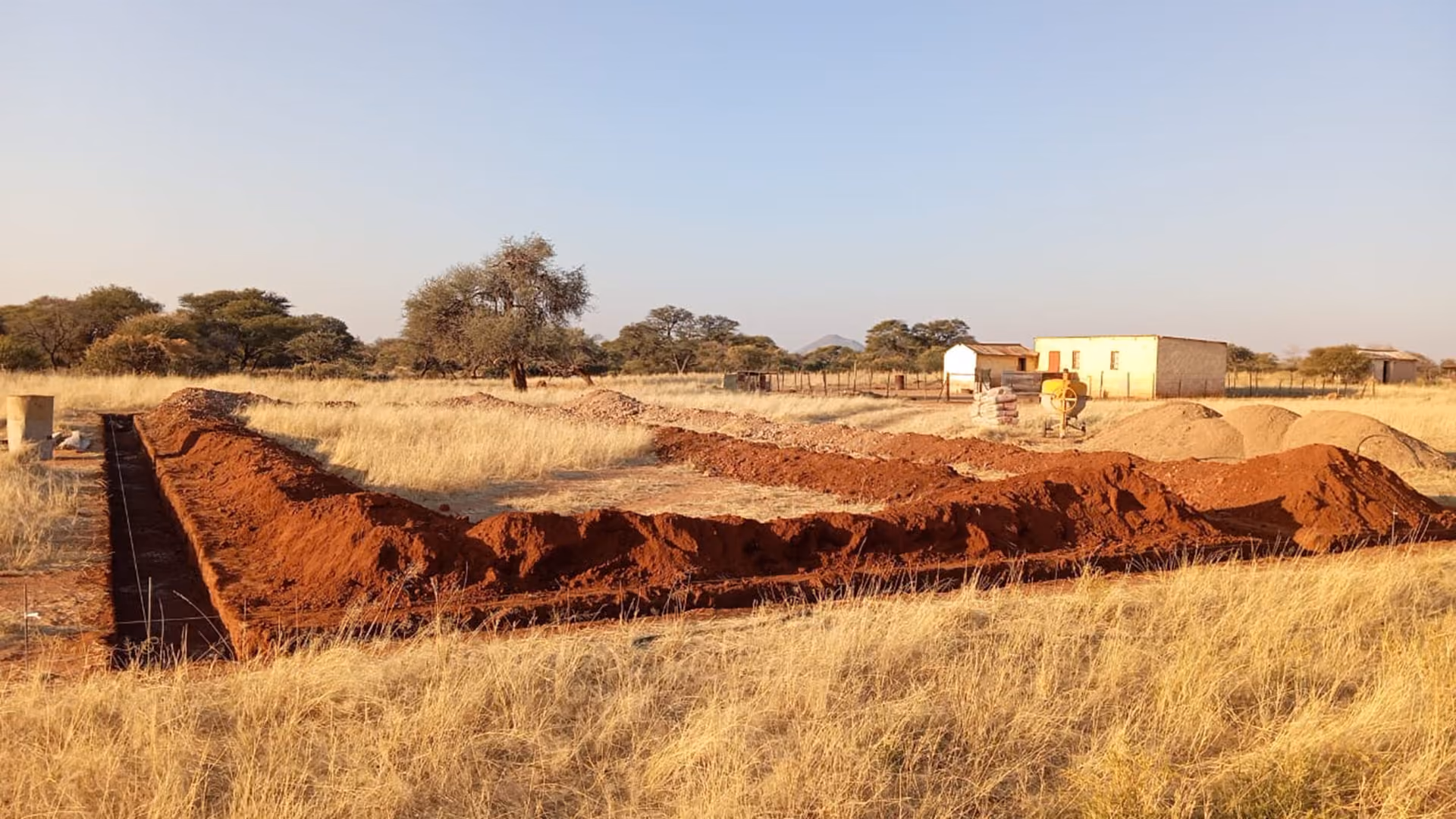 Graben einer Baustelle auf einem Feld mit Baum, Gebäuden und Baumaterial im Hintergrund.
