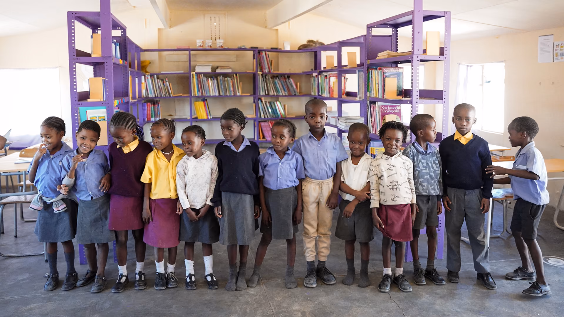Eine Gruppe von Schulkindern in Uniform steht in einer Bibliothek vor lila Bücherregalen.