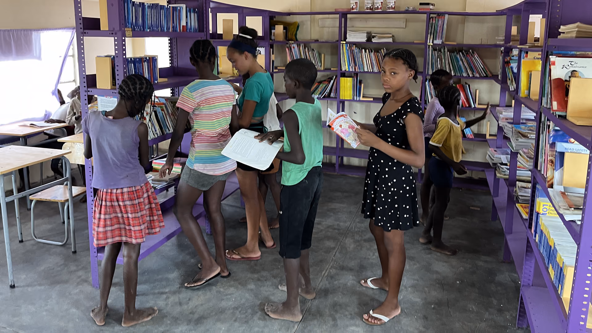 Kinder wählen Bücher aus lila Bücherregalen in einer Schule mit Tischen und Stühlen.
