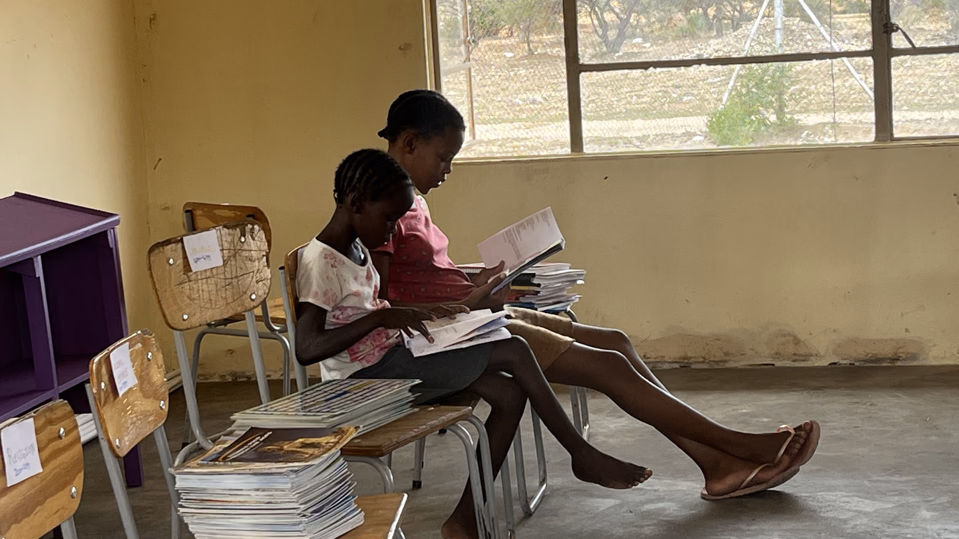 Zwei Kinder sitzen auf Stühlen und lesen Bücher in einem einfachen Raum mit Regalen und Fenster.