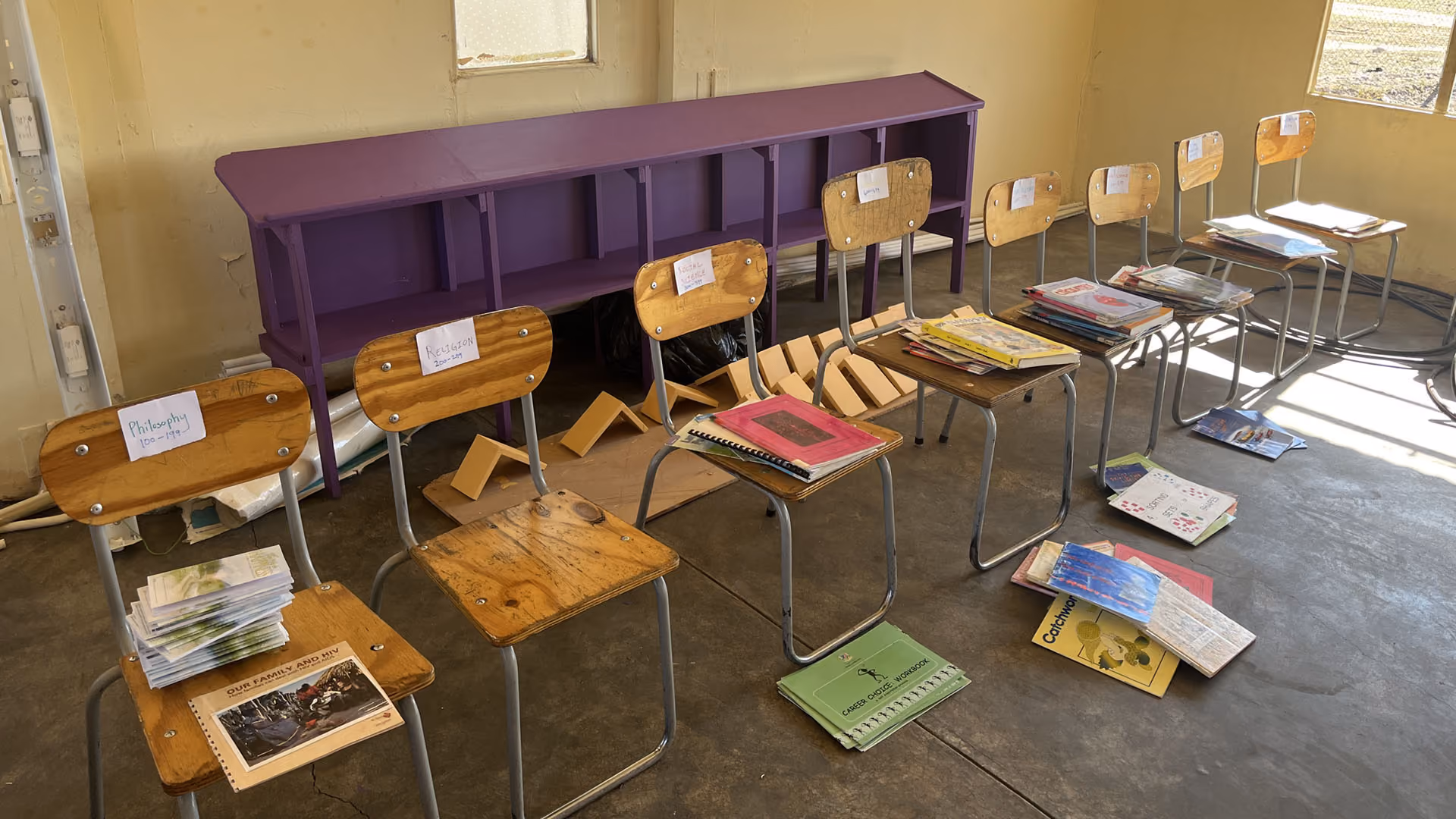 Classroom chairs labeled by subject with stacked books and booklets on seats and floor in a sunlit room.