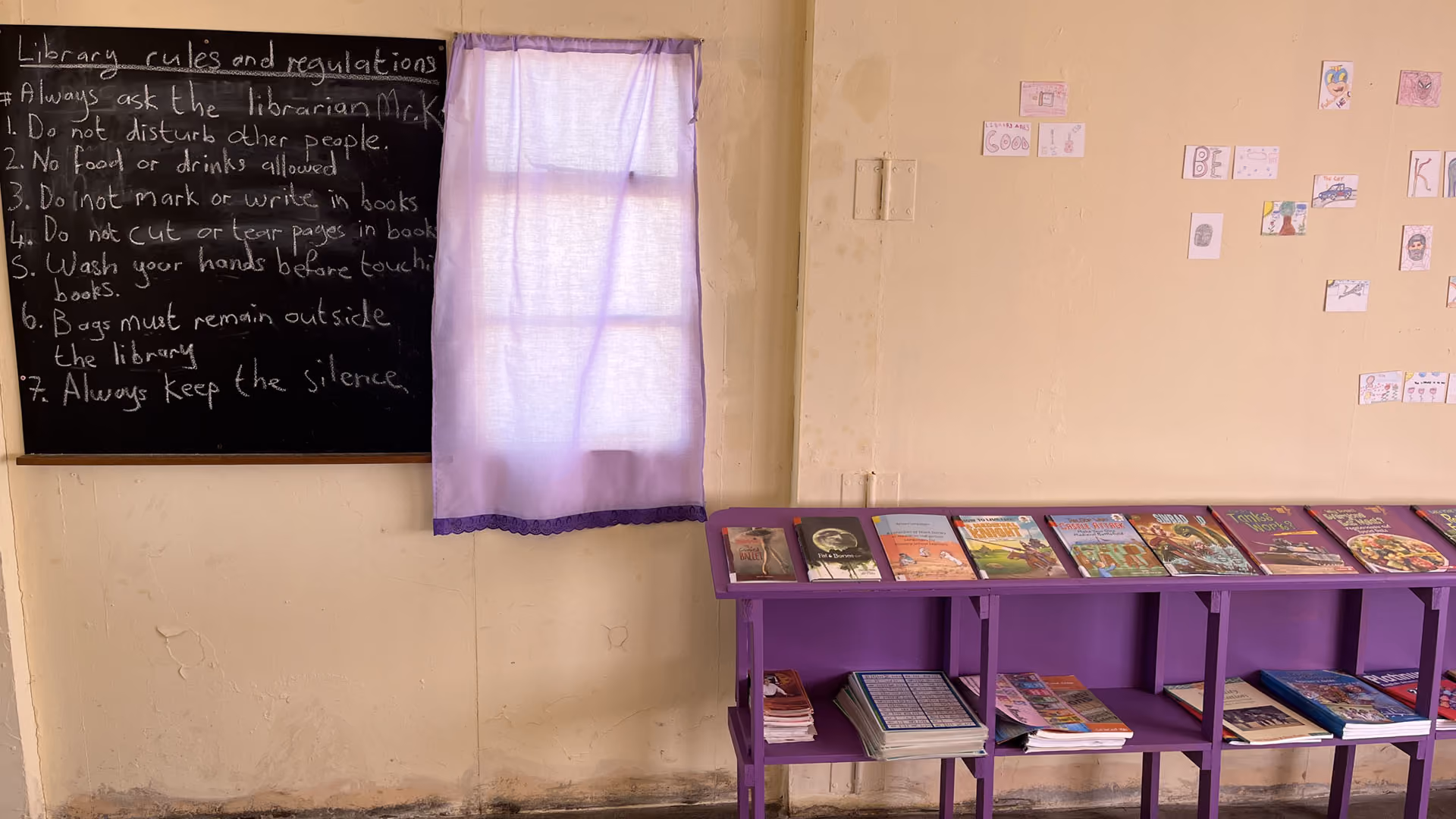 Violettes Bücherregal in einer Grundschulbibliothek neben einem Fenster und einer Tafel mit Bibliotheksregeln.