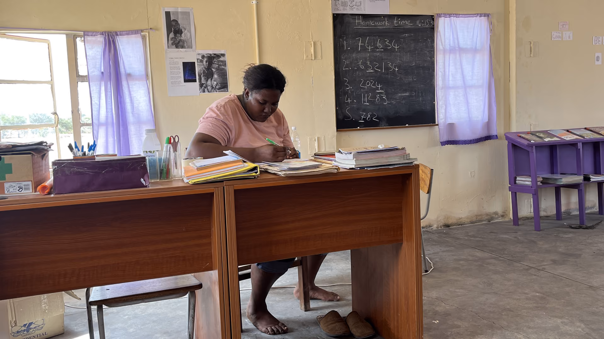 Lehrerin schreibt an Schreibtisch in einem hellen Klassenzimmer mit lila Vorhängen und Tafel.