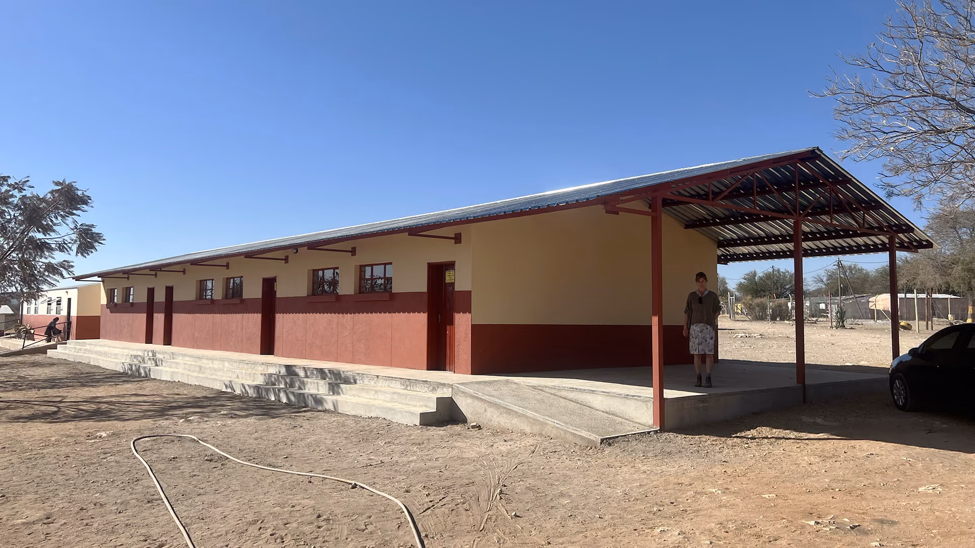 Schulgebäude mit roter und beiger Fassade, überdachtem Eingang und einer Person davor.