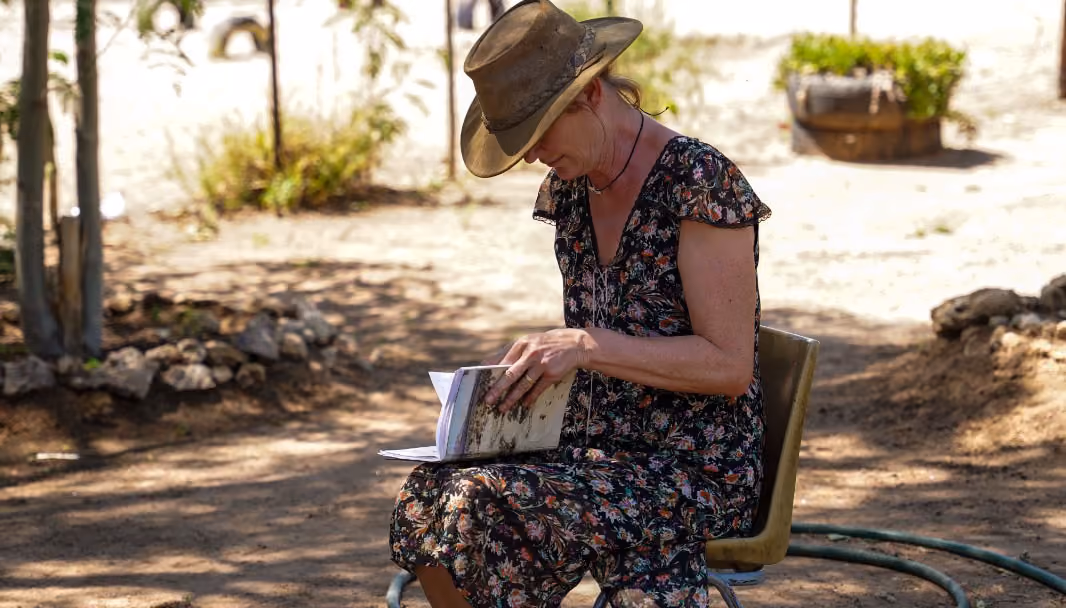 Frau mit Hut sitzt draußen auf einem Stuhl und liest ein Buch.