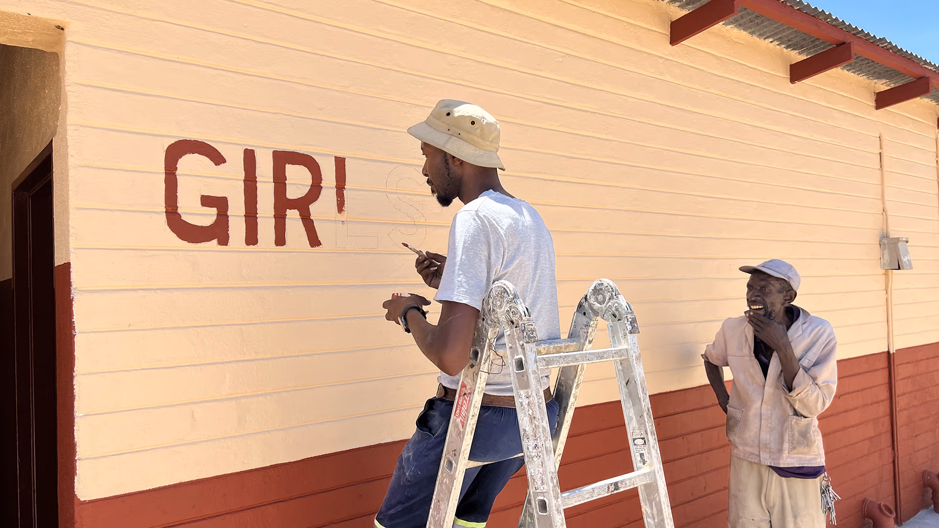 Mann auf Leiter malt ‚GIRL'S‘ auf Wand, ein älterer Mann schaut zu, vor Gebäude mit roten und gelben Wänden.