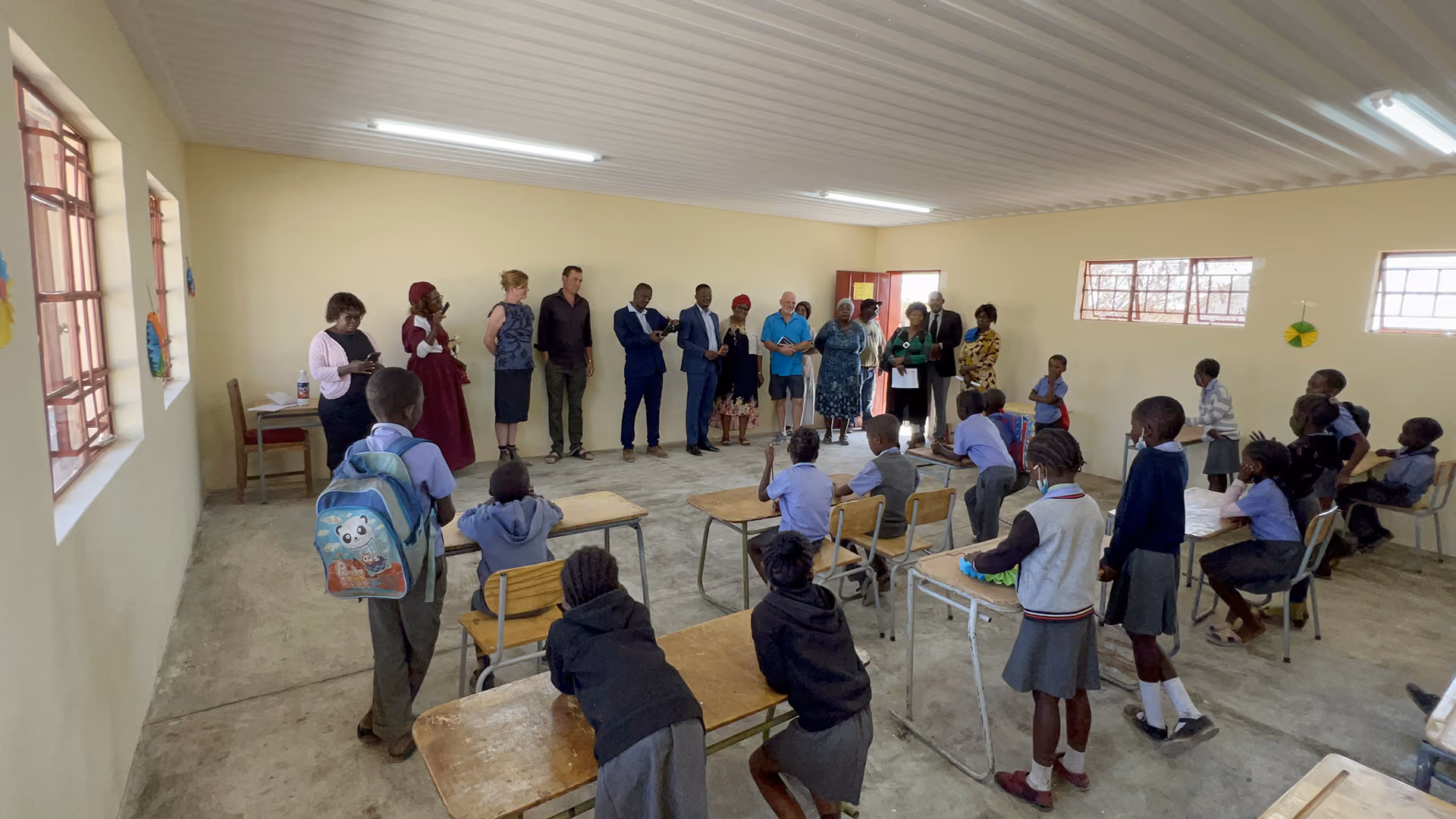 Schulkinder und Erwachsene in einem Klassenzimmer mit Holztischen und Fenstern.