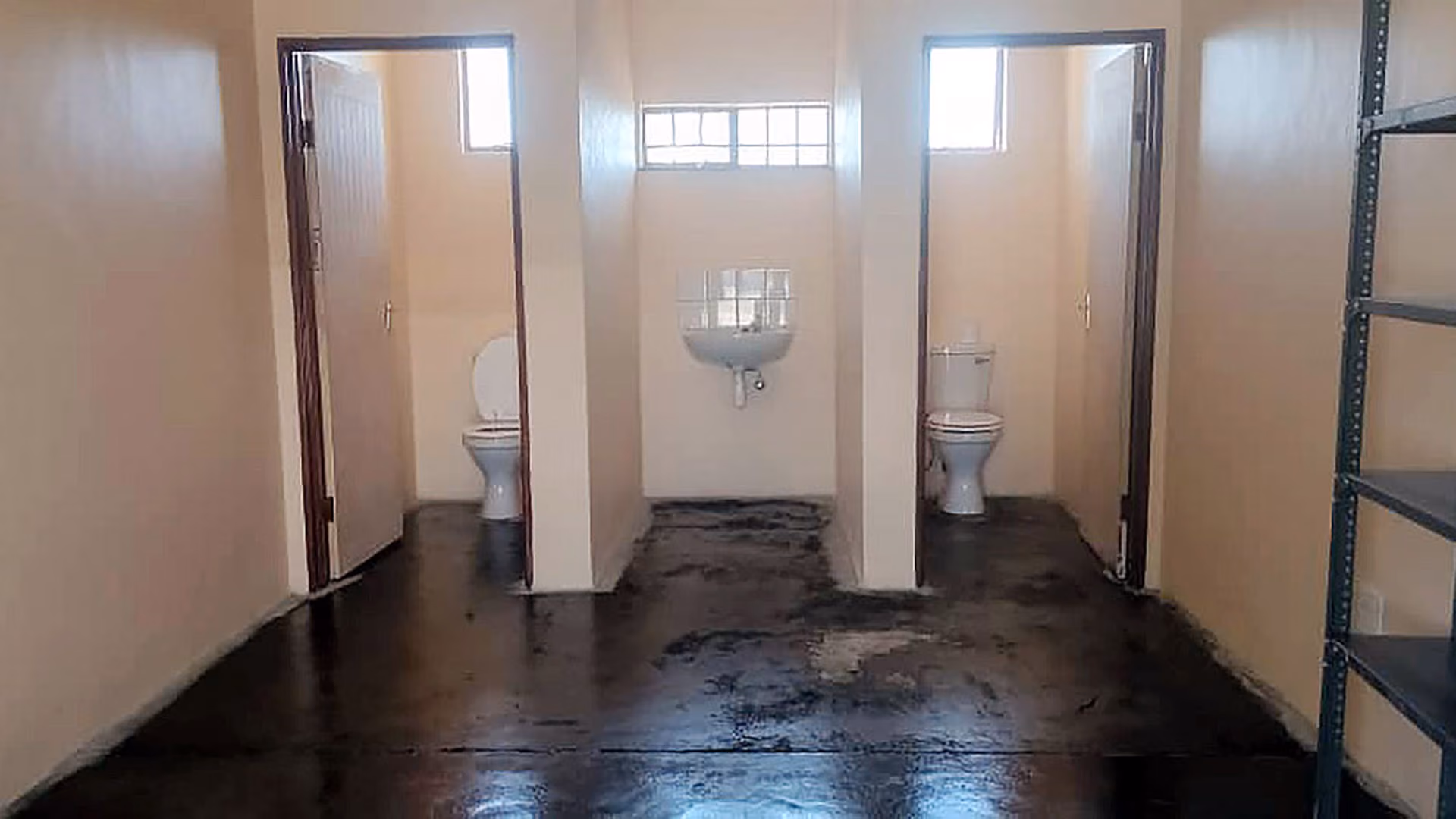 Zwei Toiletten mit jeweils geöffneter Tür und ein Waschbecken dazwischen in einem Raum.