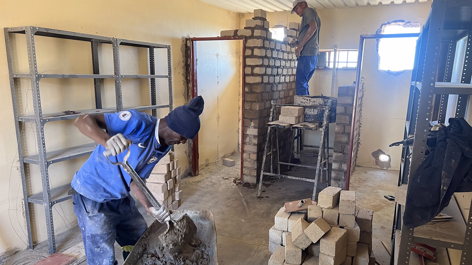 Zwei Männer bauen eine Innenwand aus Ziegelsteinen in einem Raum mit Regalen auf beiden Seiten.