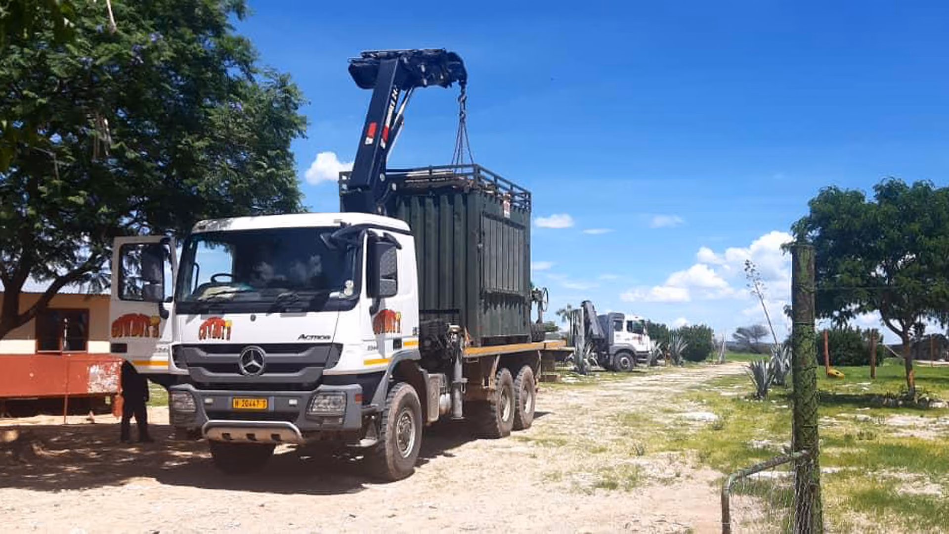 Lastwagen mit Kran hebt großen grünen Container bei sonnigem Himmel auf unbefestigtem Gelände.