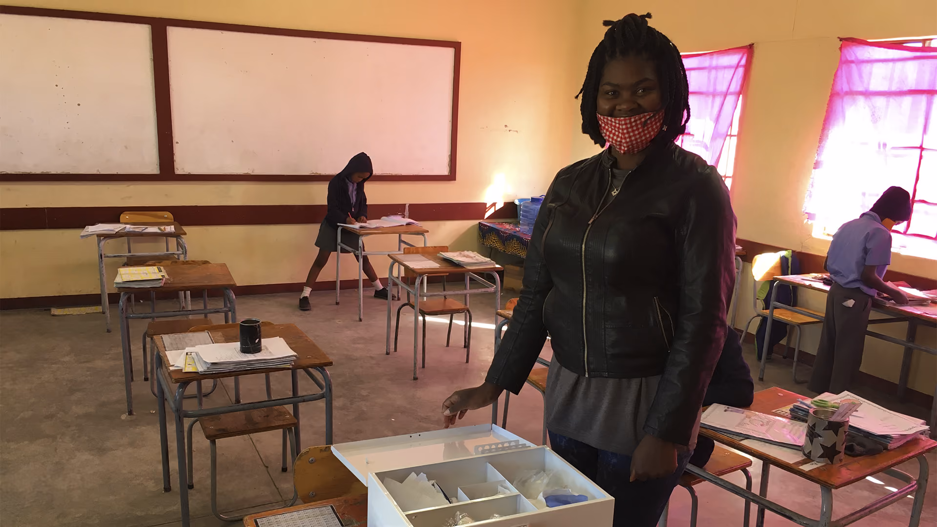 Frau mit roter Maske in Klassenzimmer mit Kindern und Tischen, einige Kinder schreiben an Pulten.
