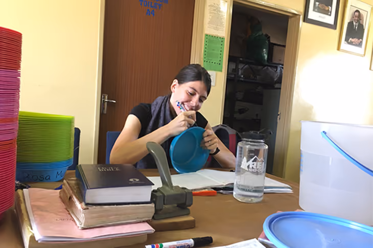 Frau beschriftet einen blauen Plastikbehälter in einem Raum mit Lehrmaterialien und Wasserflasche.