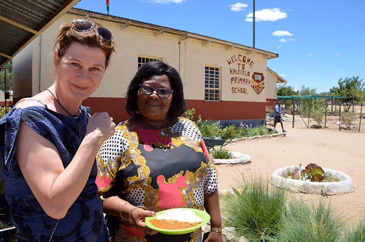 Two women stand outside a primary school; one holds a plate of food with a school building in the background.