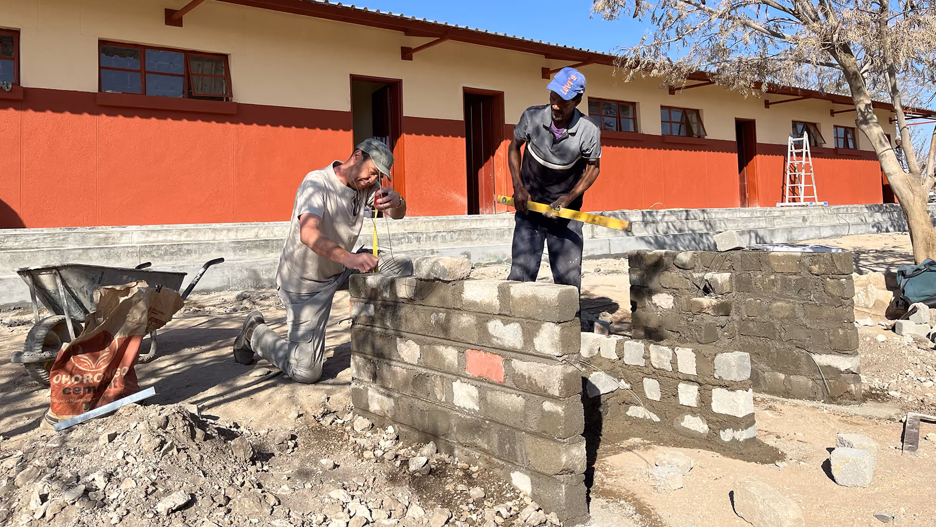 Zwei Männer messen und überprüfen eine Mauer aus Betonblöcken vor einem roten Gebäude.