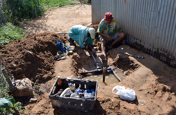 Zwei Männer reparieren Wasserleitungen in einer Erdgrube neben einem Wellblechgebäude, Werkzeugkasten daneben.